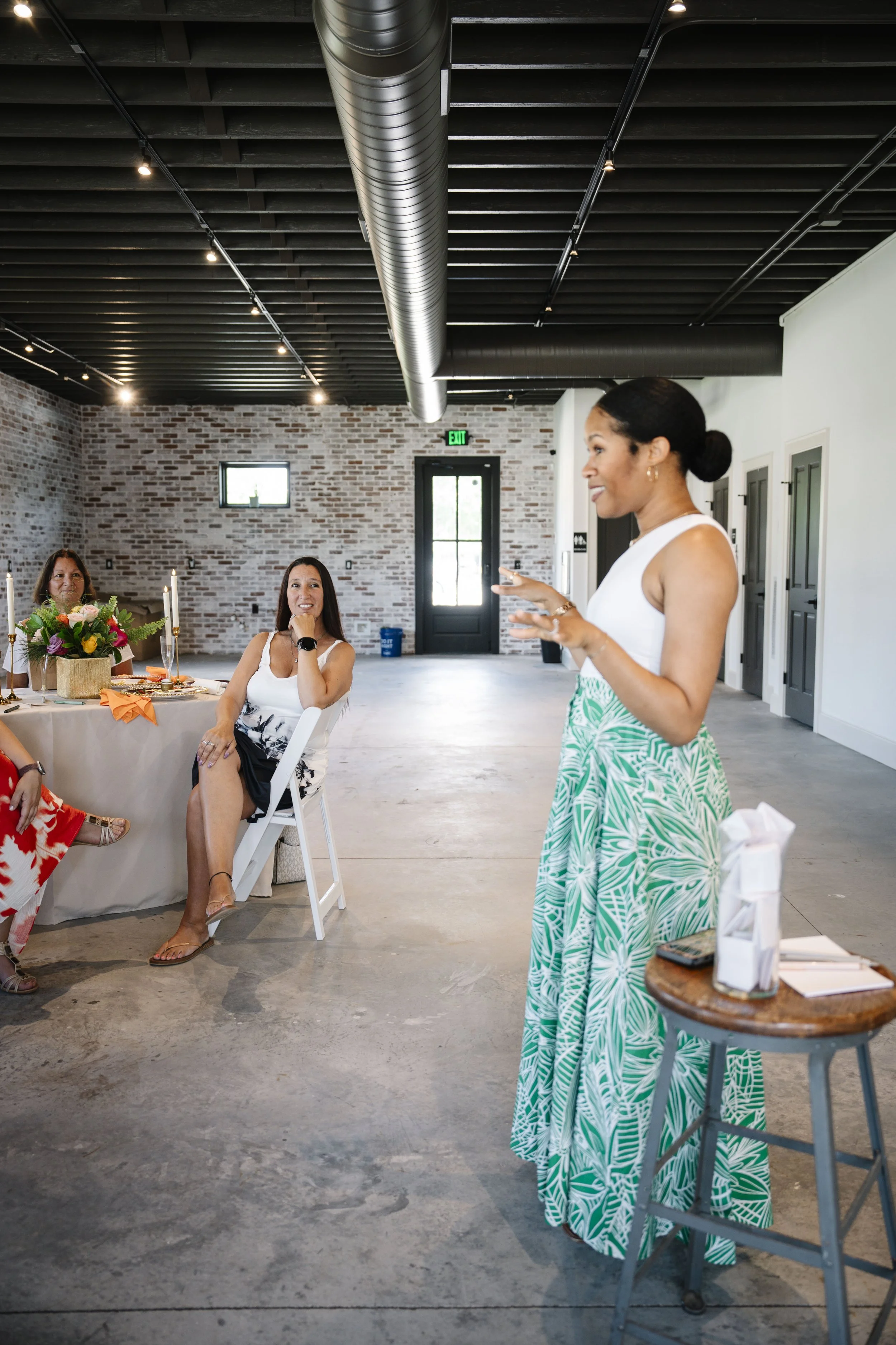 Women gathered at a celebration, sitting at a table with a floral centerpiece, candles, and decorations. One woman is speaking, and others are listening, in a room with brick walls and industrial ceiling. The women are dressed in summery clothing.