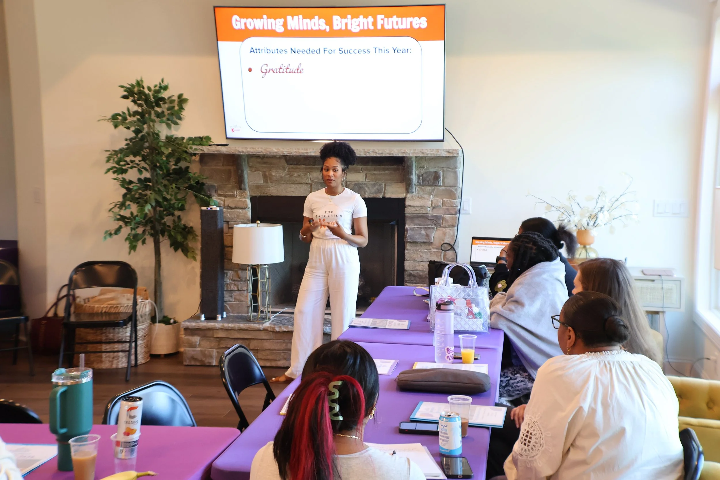 Keri Beach, Founder of The Gathering Koncept standing of a fireplace and a large screen displaying a presentation titled 'Growing Minds, Bright Futures'. She is facilitating a team retreat who's goals are to bring staff members together.