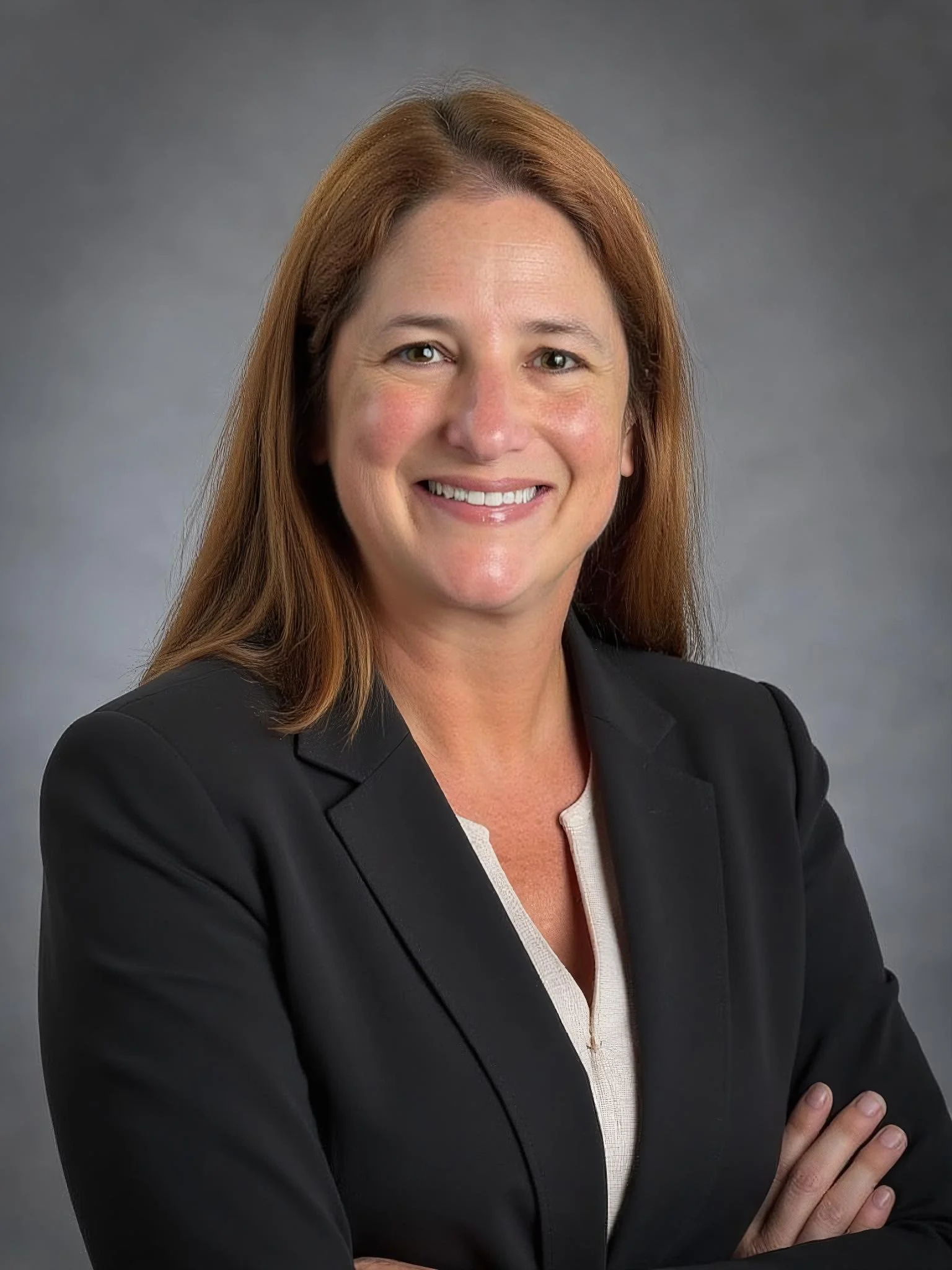 Professional woman with long red hair wearing a black blazer and a cream blouse, smiling with arms crossed against a gray studio background.