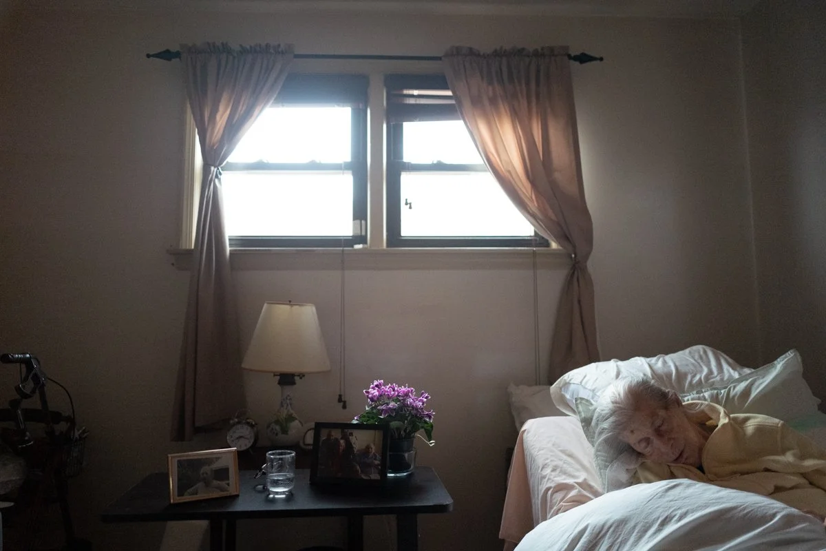 My mother lies in her bed, purple flowers to the left on a table under the window glow a little. She tilts forward under the sheets, many pillows behind her head.
