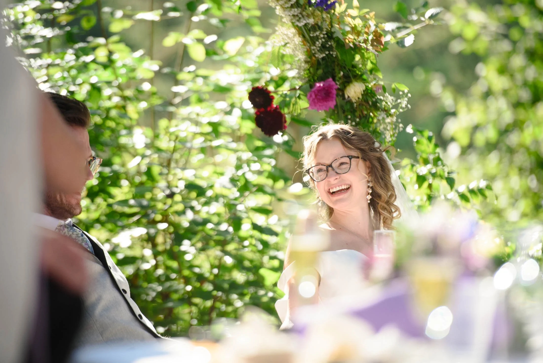 A smiling woman with glasses and curly hair, dressed in white, sitting outdoors during a sunny wedding celebration with lush green foliage and flowers in the background at Hoyt Arboretum in Portland.