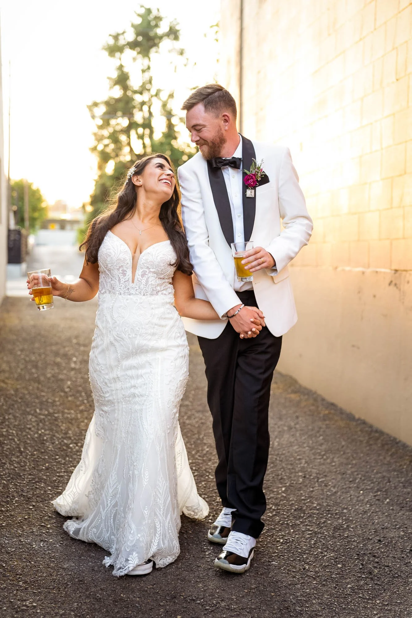A newlywed couple walking outside, holding hands and smiling at each other, both holding drinks. The bride is wearing a lace wedding gown, and the groom is in a white tuxedo jacket with a black bow tie and sneakers at Abernethy Center Ballroom.
