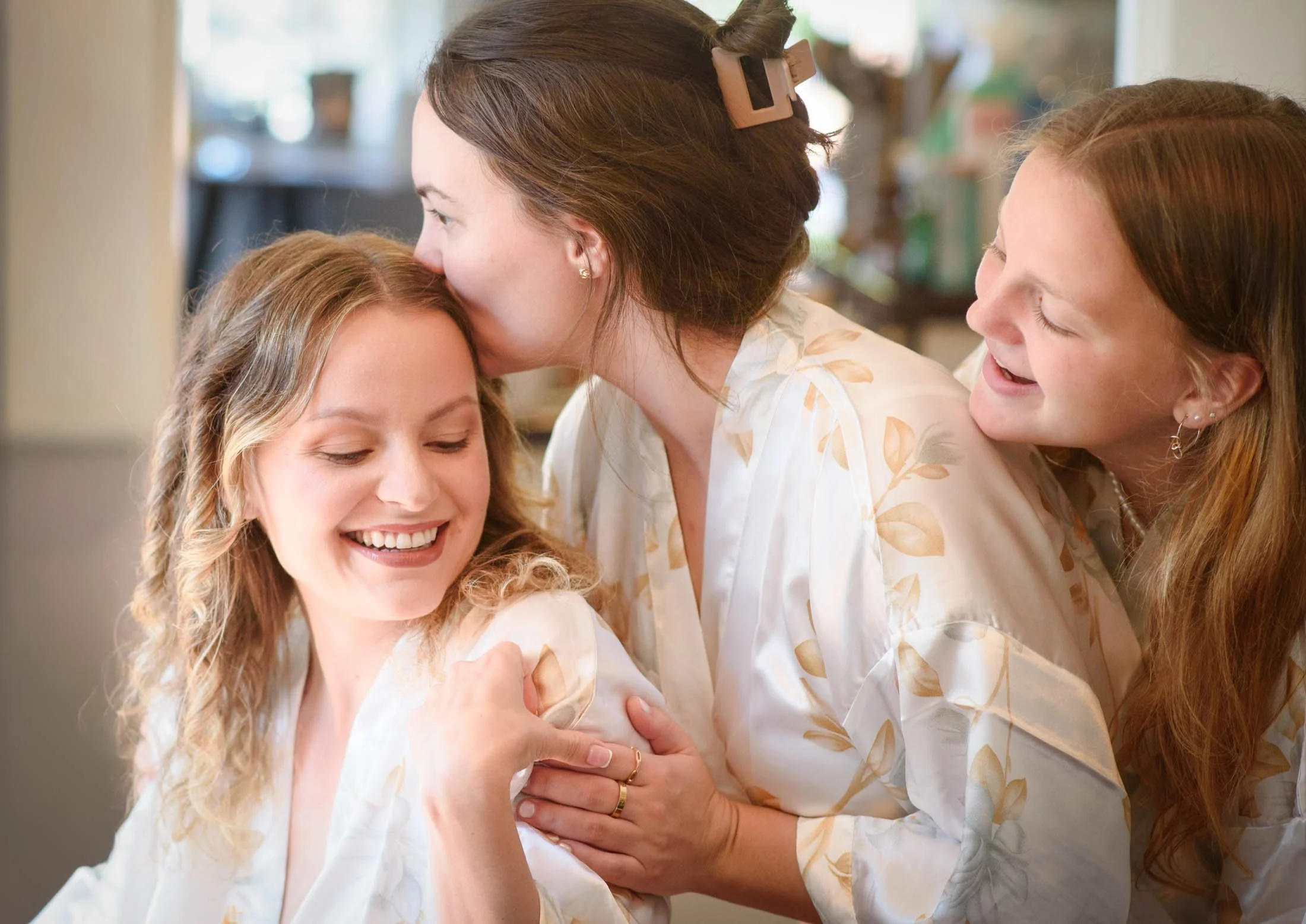 Wedding photo at Hoyt Arboretum of Three women in matching robes sharing a joyful moment, one woman being kissed on the forehead by another, while the third woman laughs and touches her shoulder.