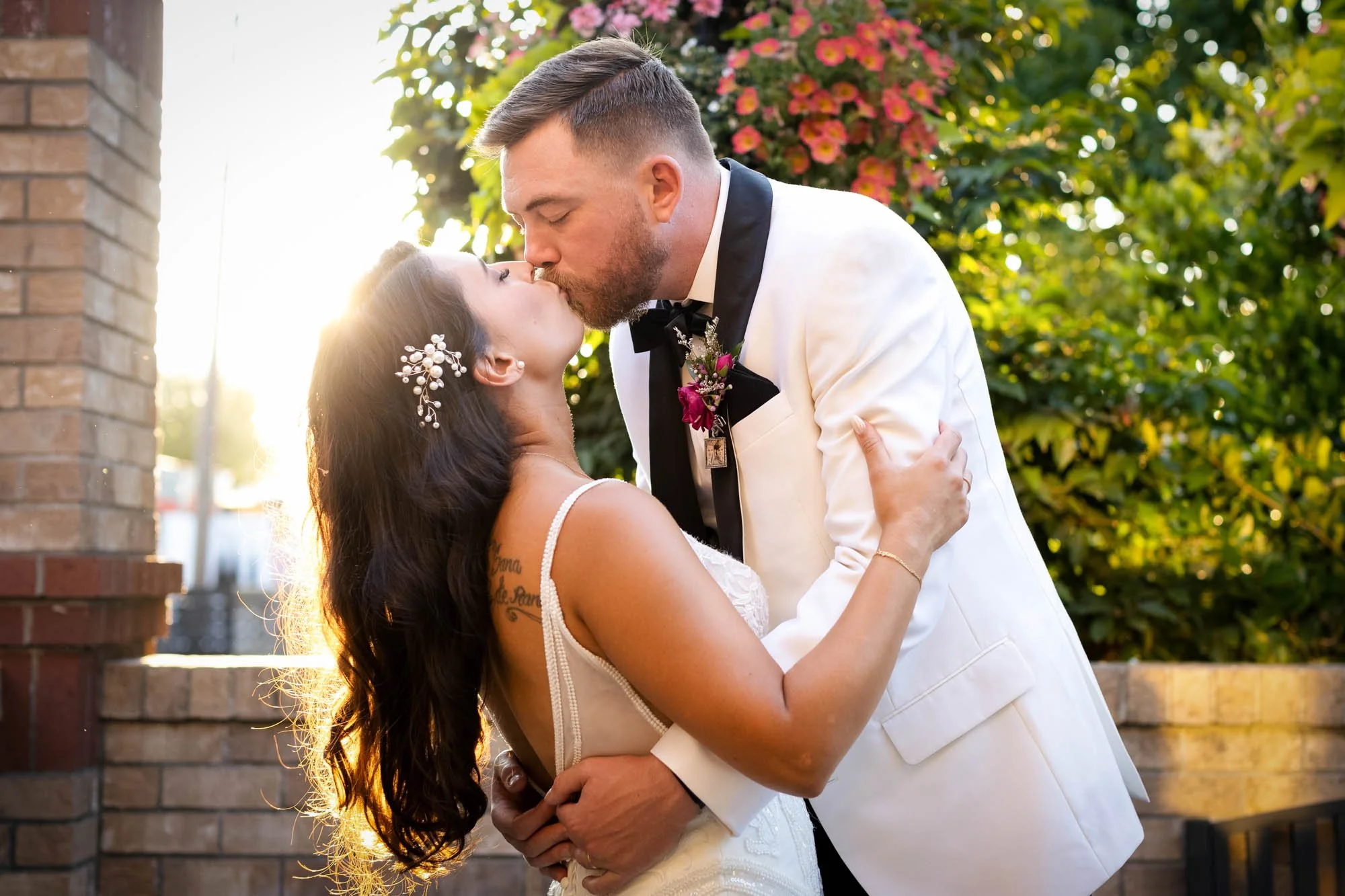A couple in wedding attire sharing a kiss outdoors with sunlight shining through trees in the background at Abernethy Center Veiled Garden and Ballroom.