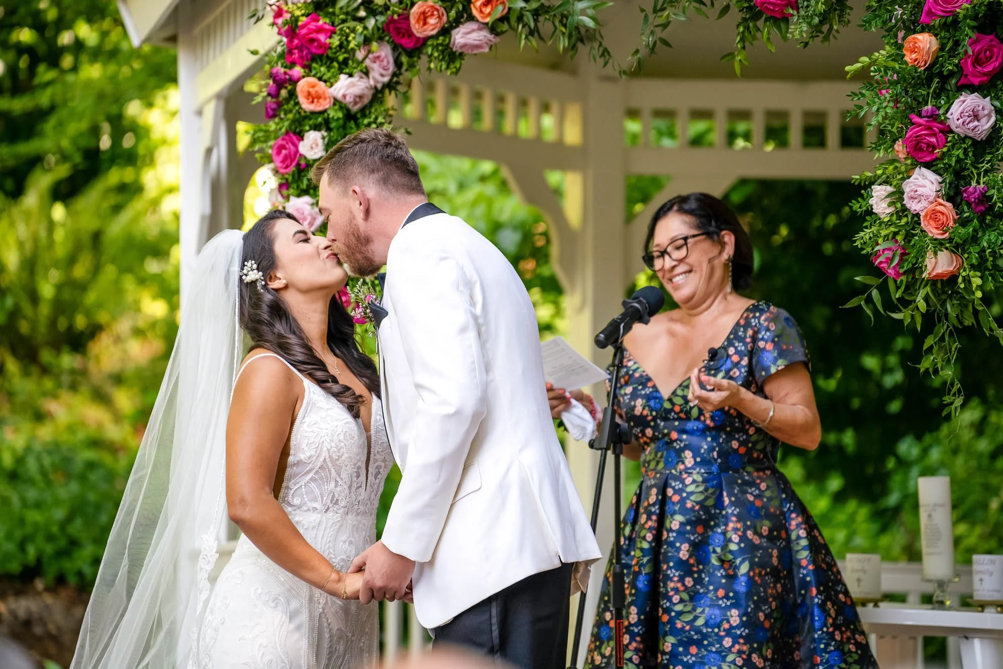 A couple getting married outdoors at Abernethy Center Abigails Garden, kissing under a floral arch, with a woman officiant smiling nearby.