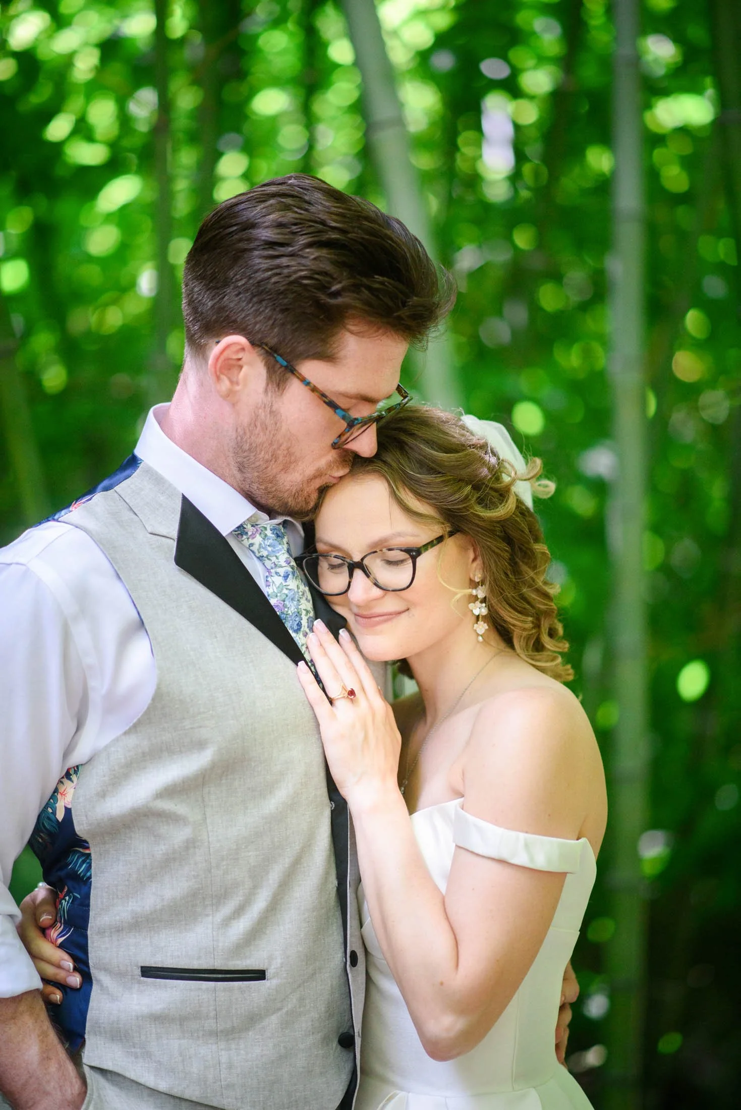 A couple in wedding attire sharing an intimate moment in a lush green forest. The man is leaning down, touching foreheads with the woman, who has her eyes closed, smiling softly  at Hoyt Arboretum in Portland.