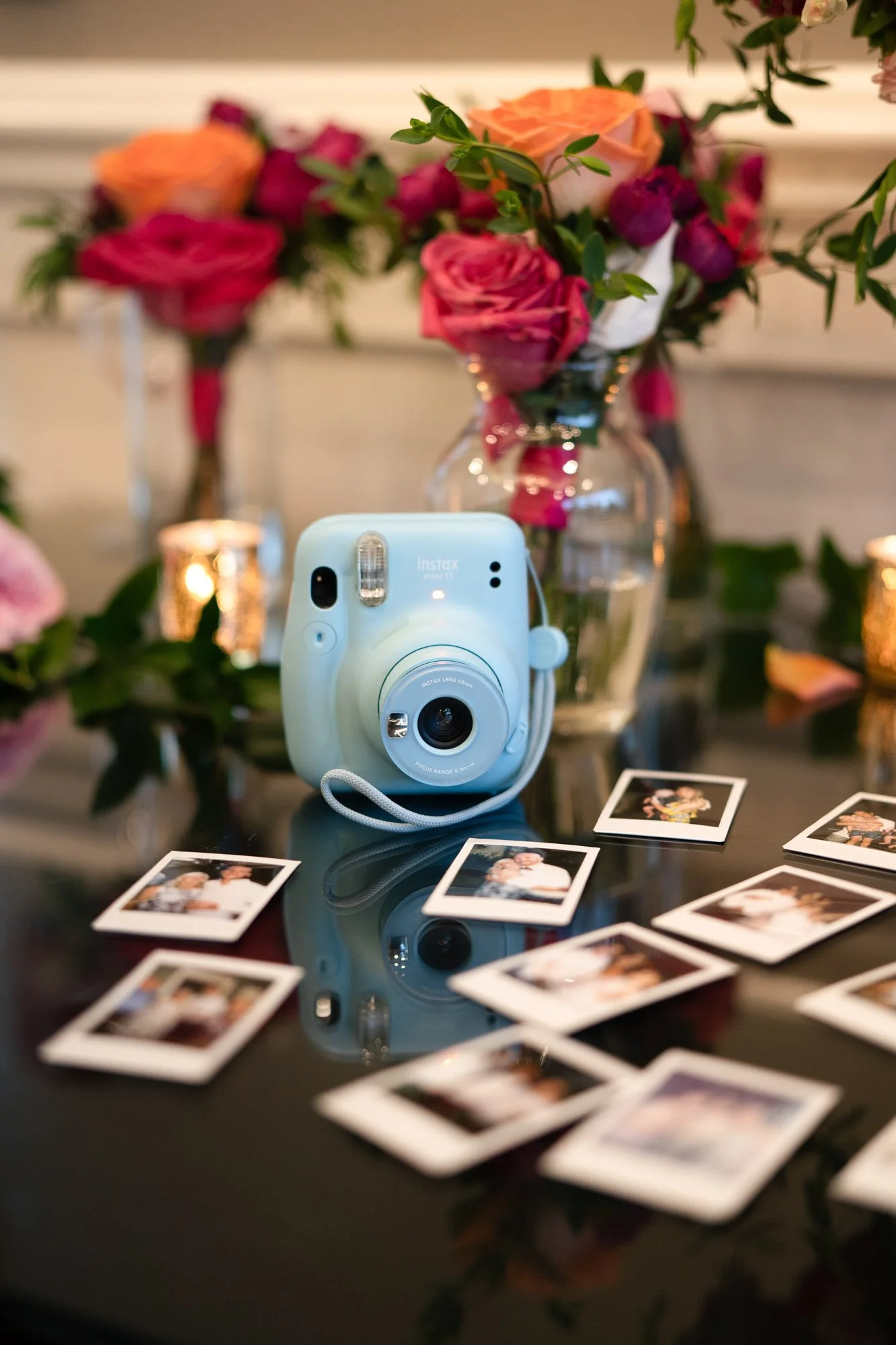 A blue instant camera on a shiny black table, surrounded by scattered instant photos, with a large vase of pink, orange, and purple roses in the background and small lit candles.