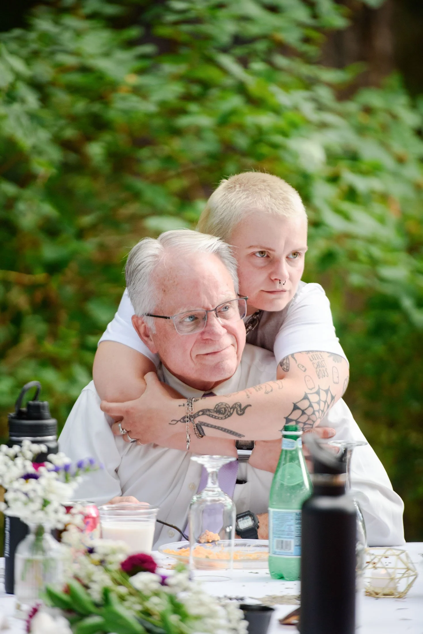 A young person with short blond hair and tattoos on their arm hugging an older man with gray hair and glasses at a table outdoors surrounded by greenery.