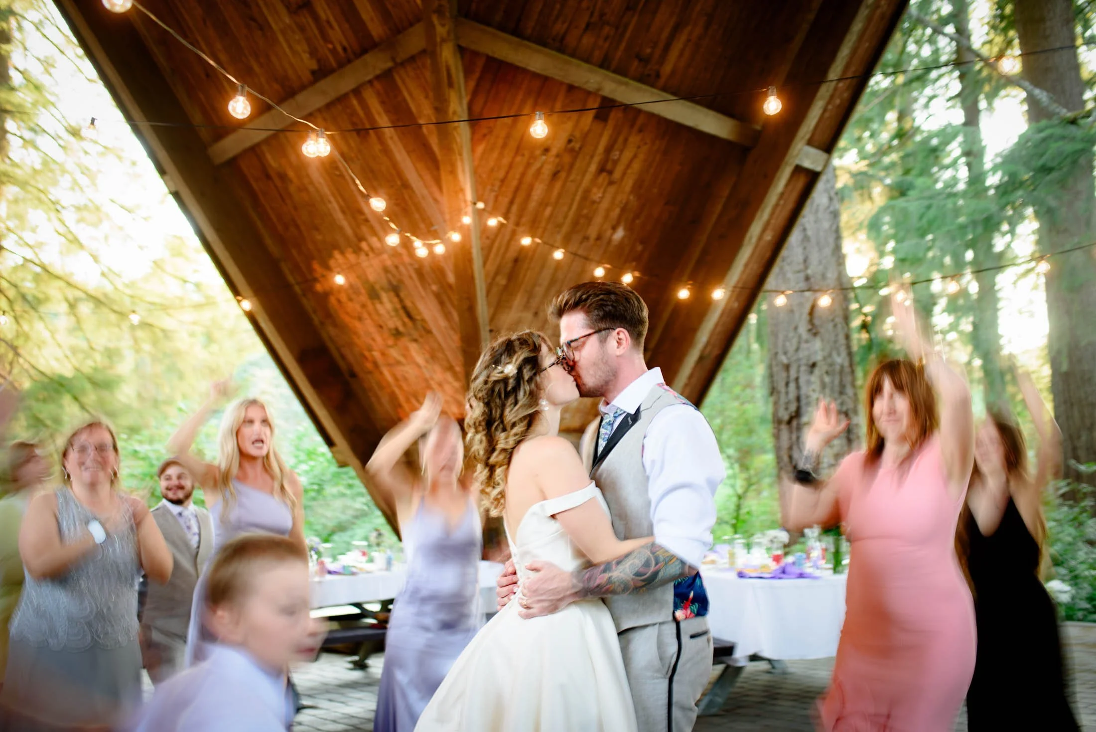 Wedding photograph at Portland's Hoyt Arboretum of A couple is kissing during a wedding reception in an outdoor pavilion, surrounded by friends and family dancing and celebrating.
