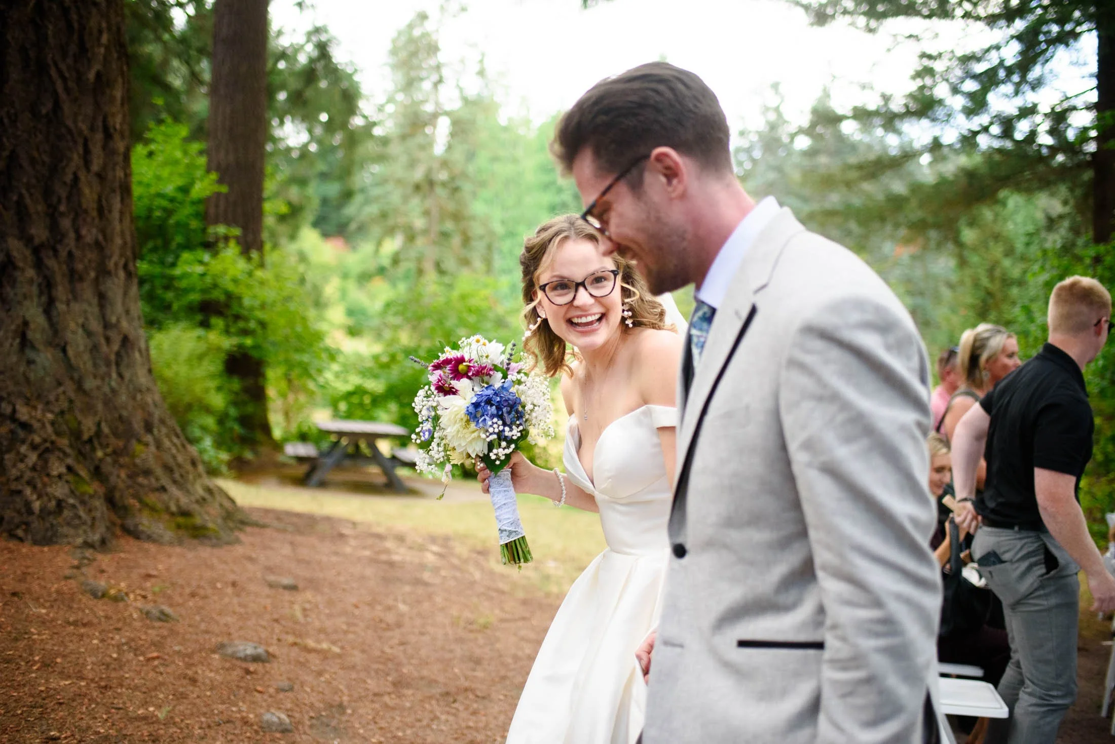 Wedding photo at Hoyt Arboretum of A bride with glasses and a bouquet laughing at her wedding outdoors, standing next to a groom in a light gray suit among trees and guests.