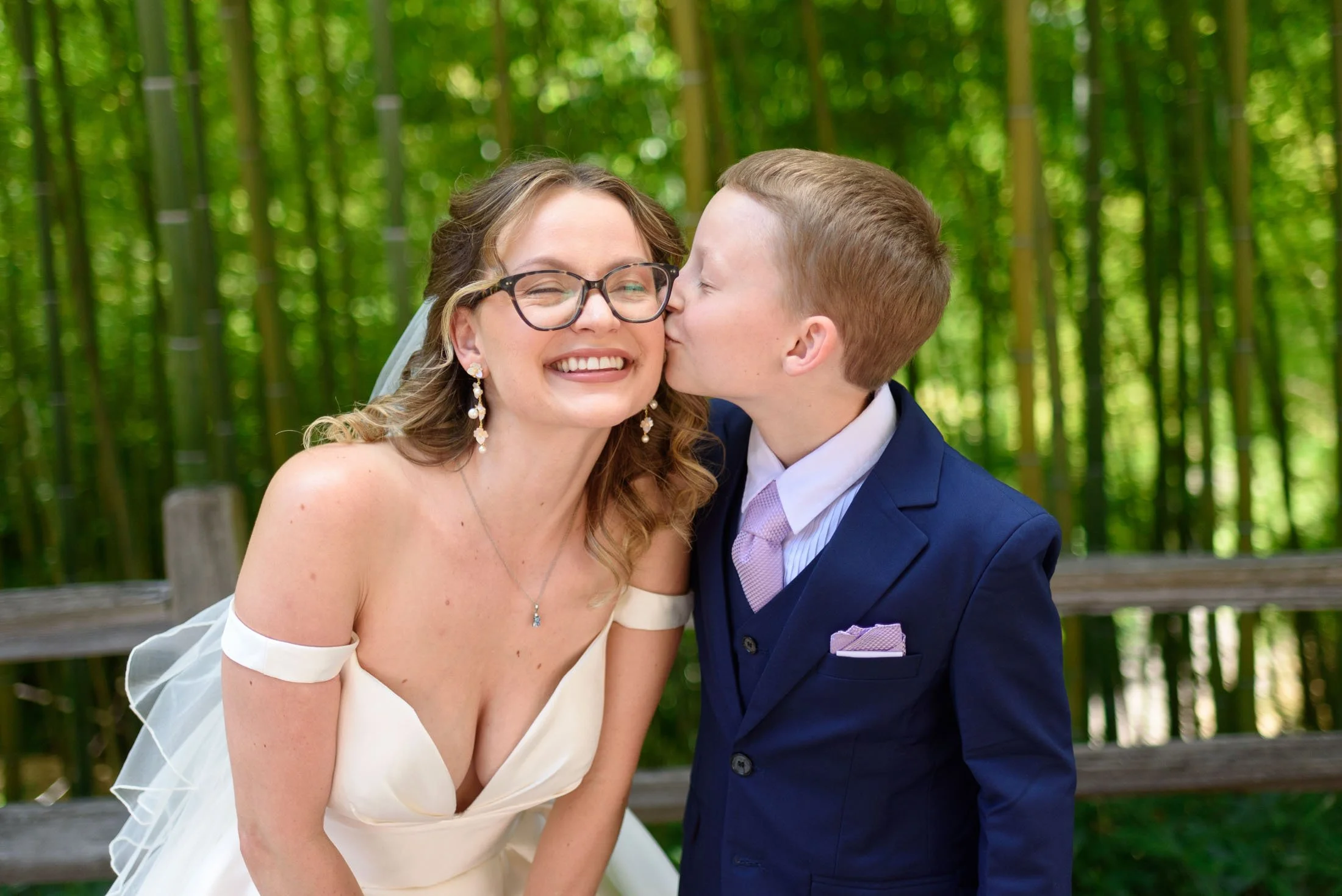 Wedding photo at Hoyt Arboretum of A bride in a wedding dress and glasses is smiling as a boy in a blue suit kisses her on the cheek in a green wooded outdoor setting.