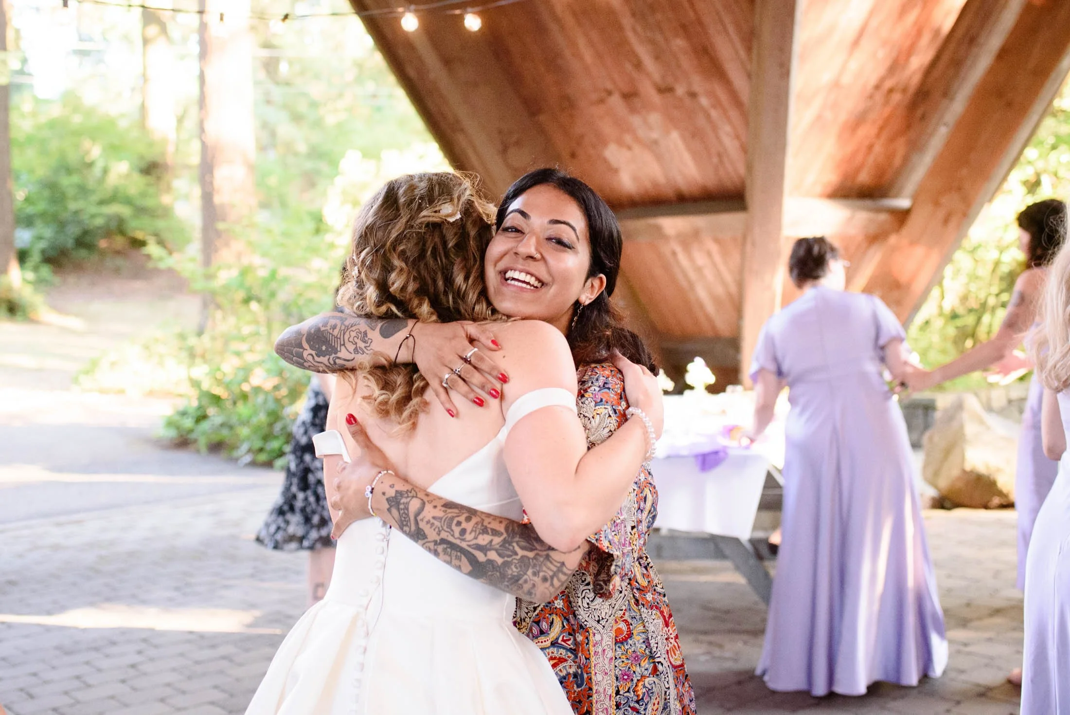 Wedding photograph at Portland's Hoyt Arboretum of Two women hugging, one in a wedding dress and the other in a colorful dress, smiling at a wedding reception outdoors under a wooden pavilion.