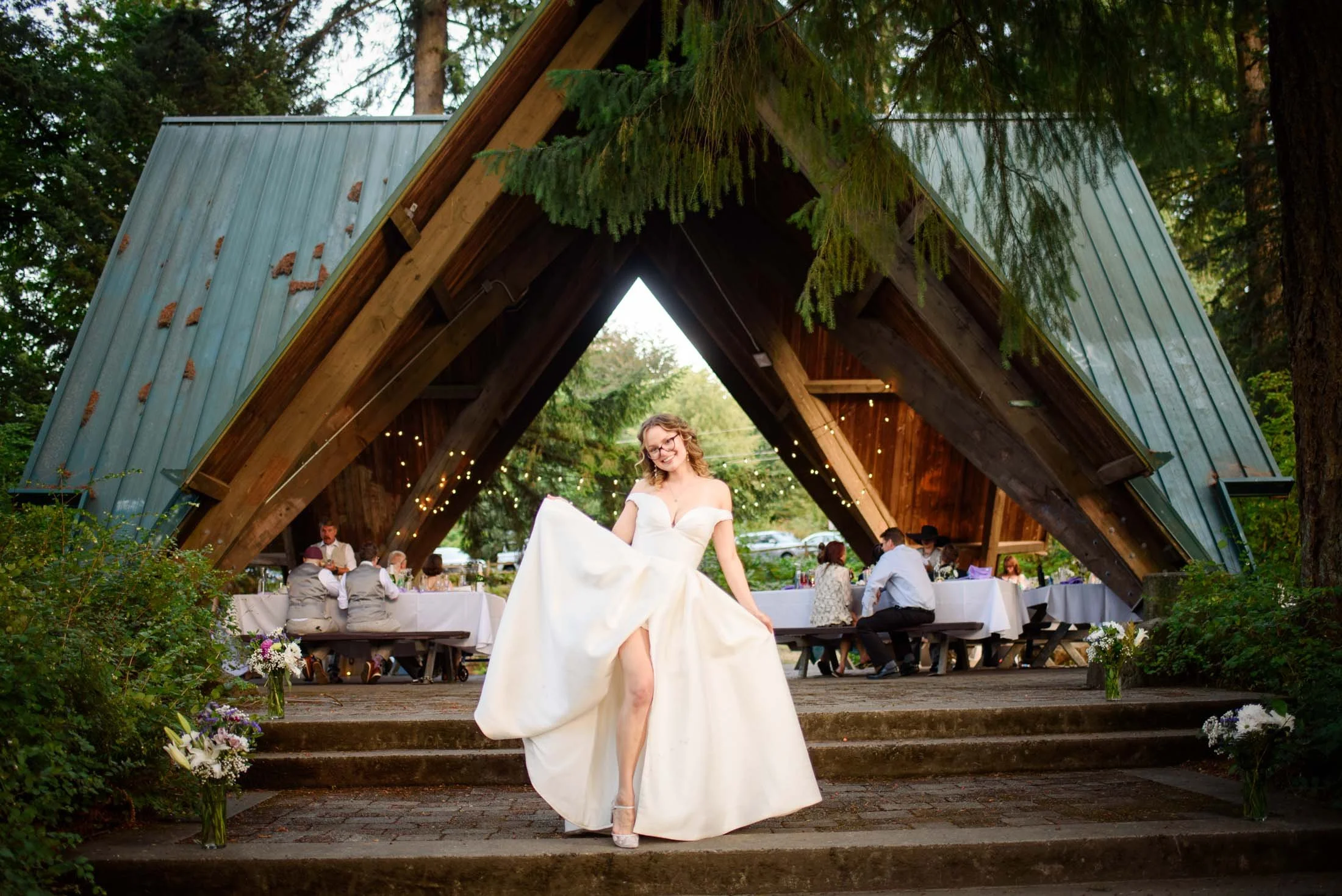 Wedding photograph at Portland's Hoyt Arboretum of A bride lifting her wedding dress in front of an outdoor wedding reception in a wooden A-frame pavilion surrounded by greenery.