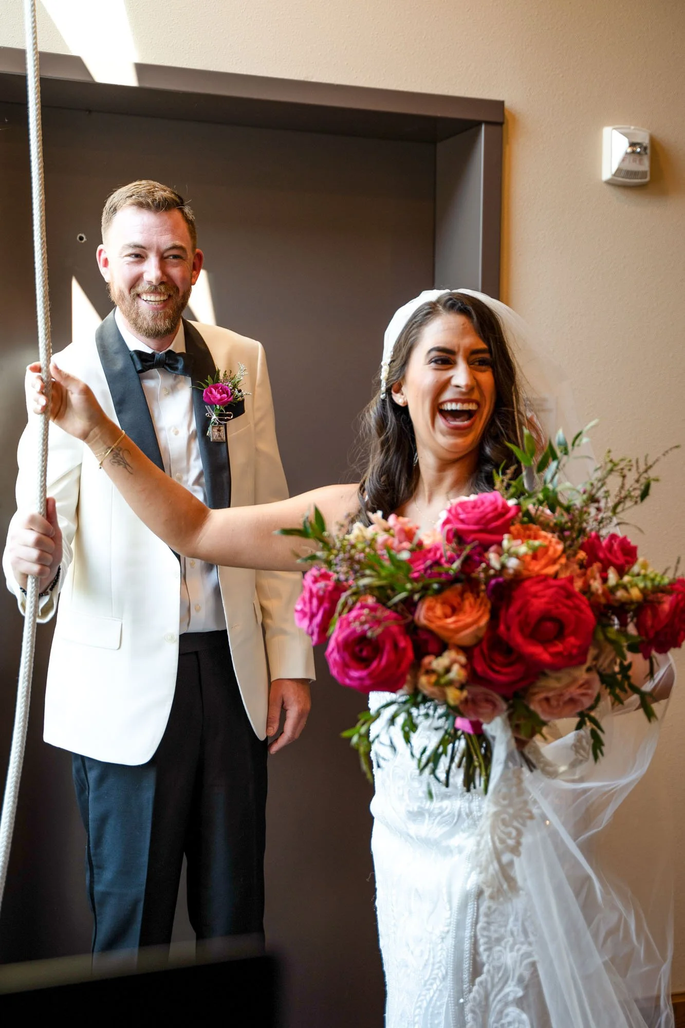 A bride in a white wedding dress holding a large bouquet of pink, coral, and peach flowers, laughing joyfully. A groom in a tuxedo with a white jacket, black bow tie, and pink flower boutonniere stands beside her, smiling.