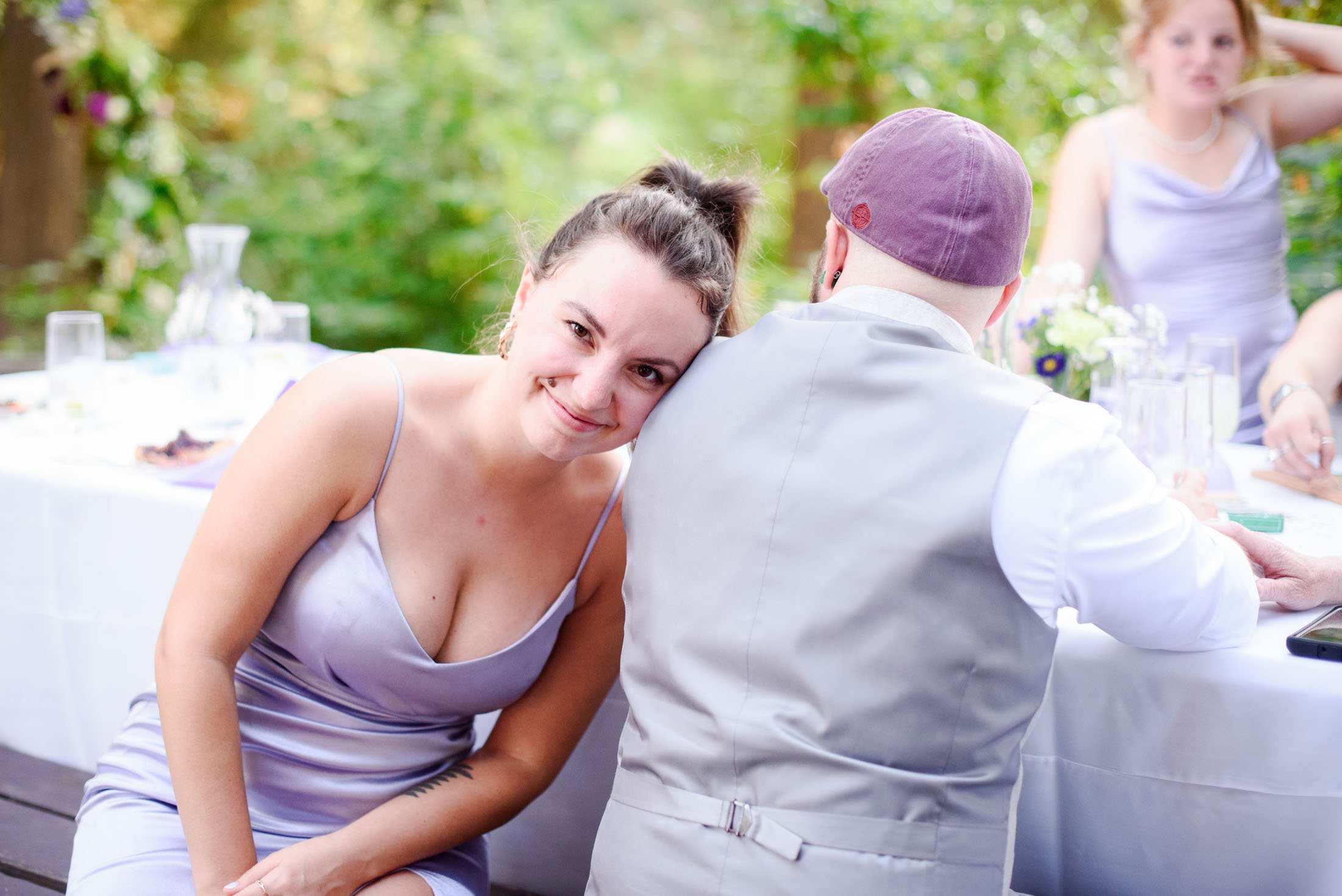 Wedding photograph at Portland's Hoyt Arboretum of A young woman with dark hair in a bun smiling and leaning her head on a person's shoulder during an outdoor gathering, with a table set with drinks and flowers in the background.