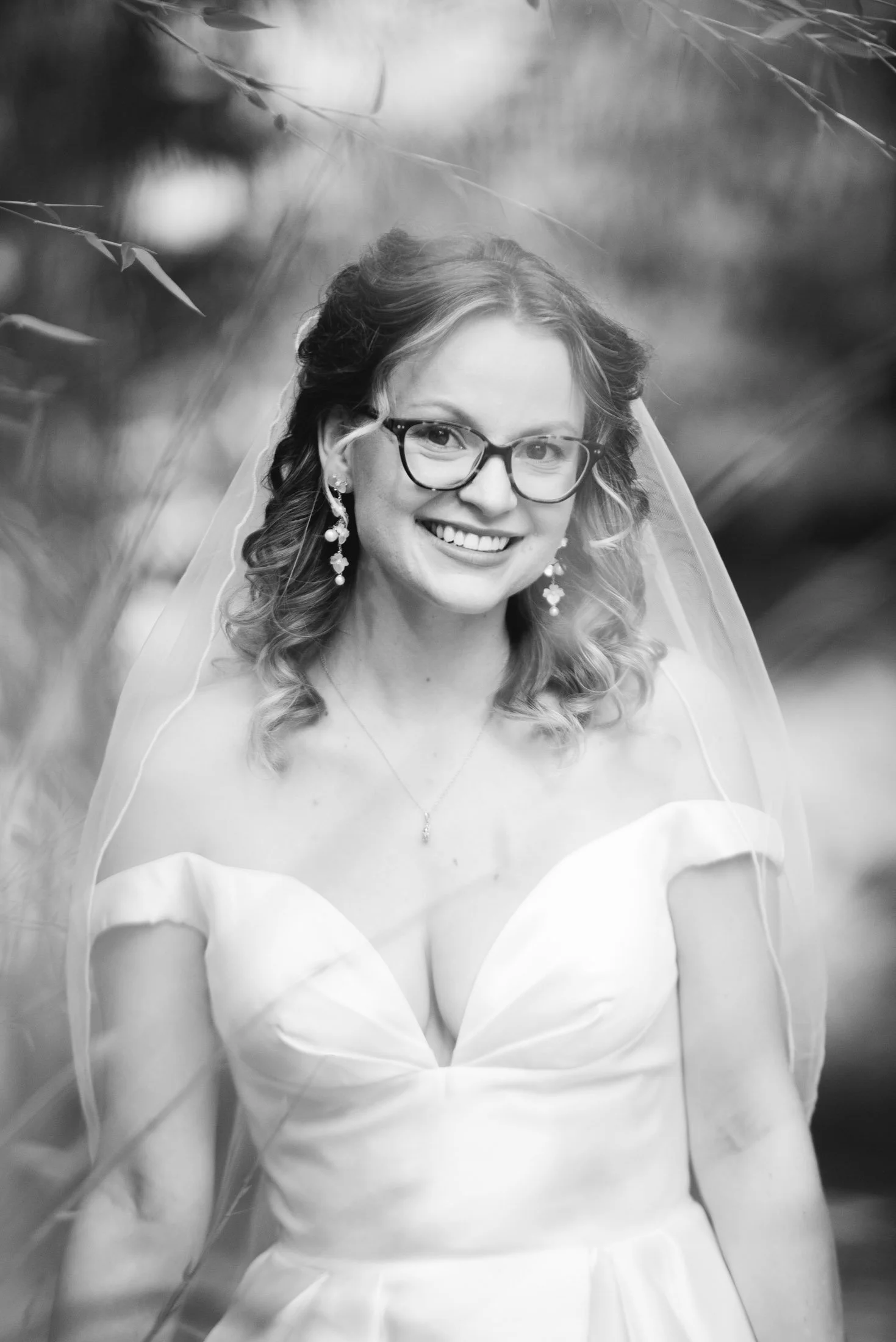 Wedding photo at Hoyt Arboretum of Black and white portrait of a woman in a wedding dress with a veil, glasses, and earrings, smiling outdoors.