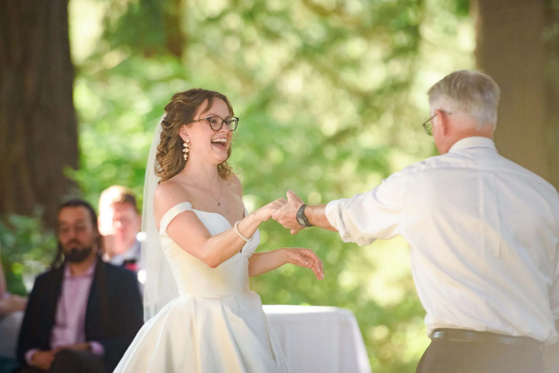 Wedding photograph at Portland's Hoyt Arboretum of A bride and an older man, possibly her father, are holding hands and smiling at each other during a wedding ceremony outdoors in a forested area. 