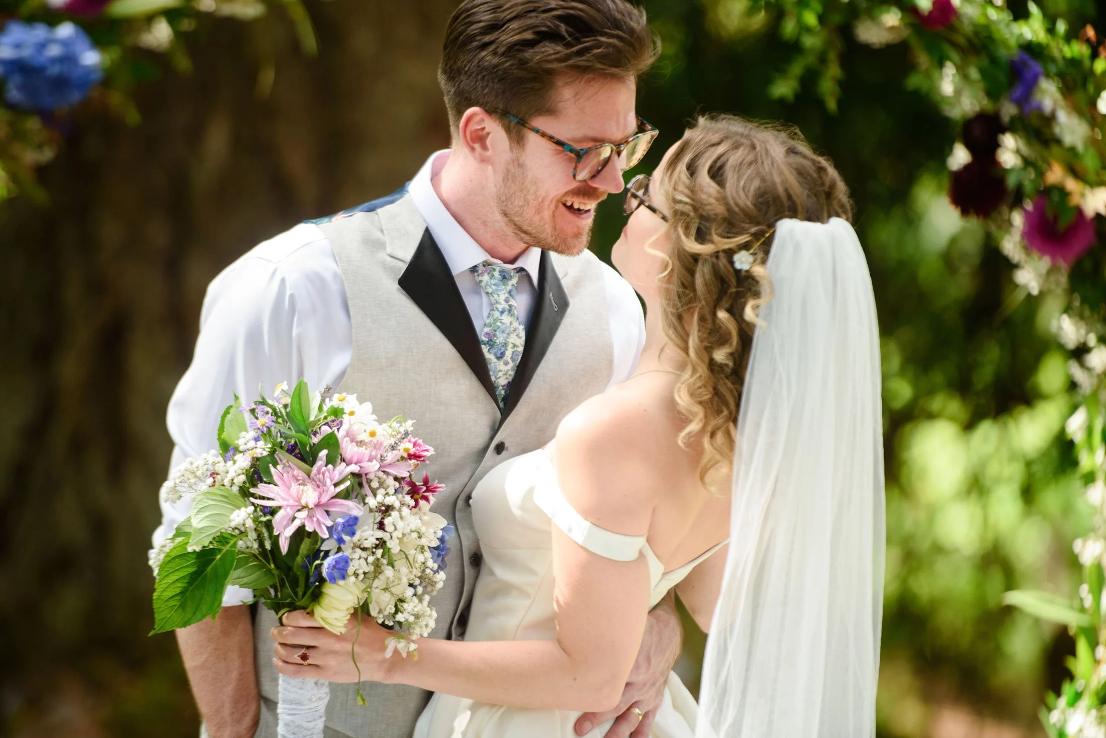 A bride and groom share a romantic moment at their wedding outdoors, with greenery and flowers in the background. The bride holds a colorful bouquet, and both are smiling and looking at each other at Hoyt Arboretum in Portland.