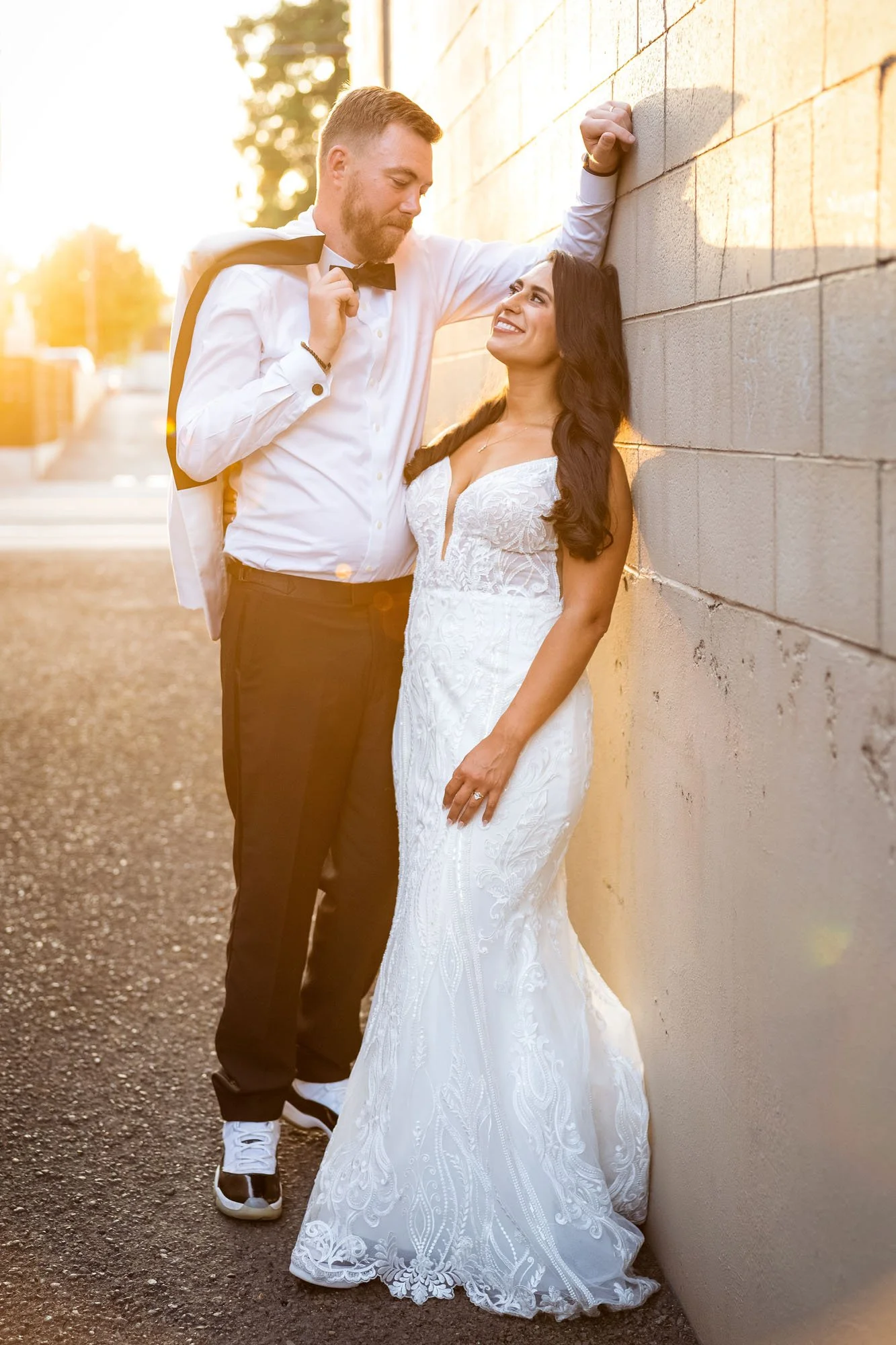A couple dressed in wedding attire standing outdoors during sunset, with the man leaning against a brick wall and the woman smiling up at him at Abernethy Center Veiled Garden and Ballroom.