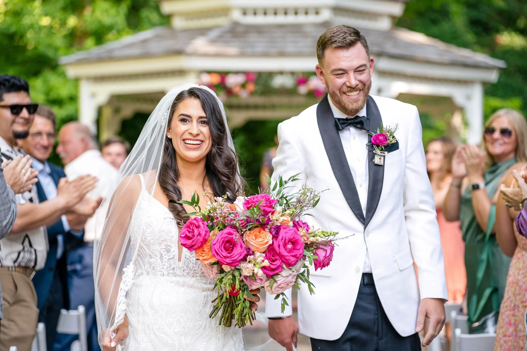 Wedding photography at Abernethy Center's Abigail's Garden of a Bride holding a bouquet of pink, orange, and purple flowers, smiling, walking with groom in white tuxedo with black lapel and bow tie, surrounded by guests clapping outdoors.