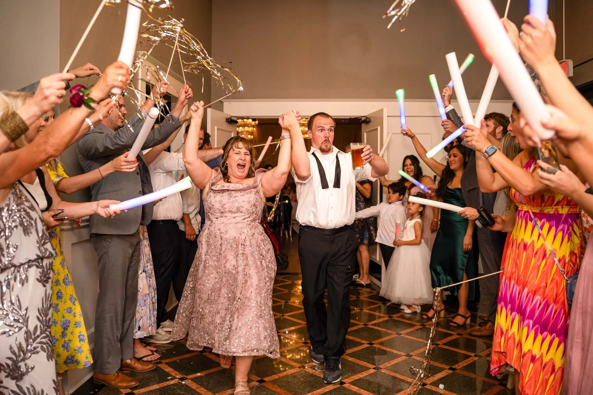 Wedding photograph at Abernethy Center Veiled Garden and Ballroom of Couple walking through a tunnel of cheering guests holding glow sticks and streamers at a celebration, likely a wedding reception.