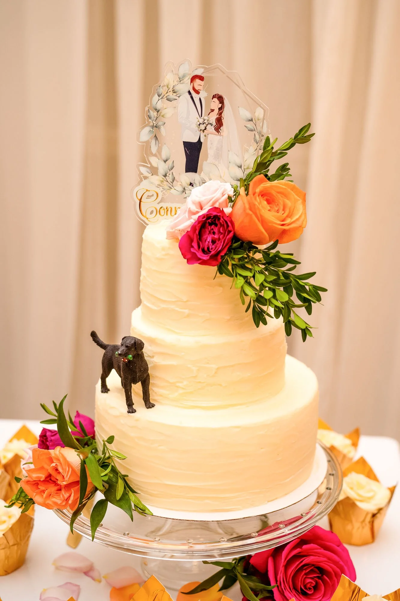 A two-tiered wedding cake decorated with fresh flowers, a dog figurine, and a custom topper of a couple in wedding attire, placed on a white table with scattered rose petals and gold foil containers in the background.