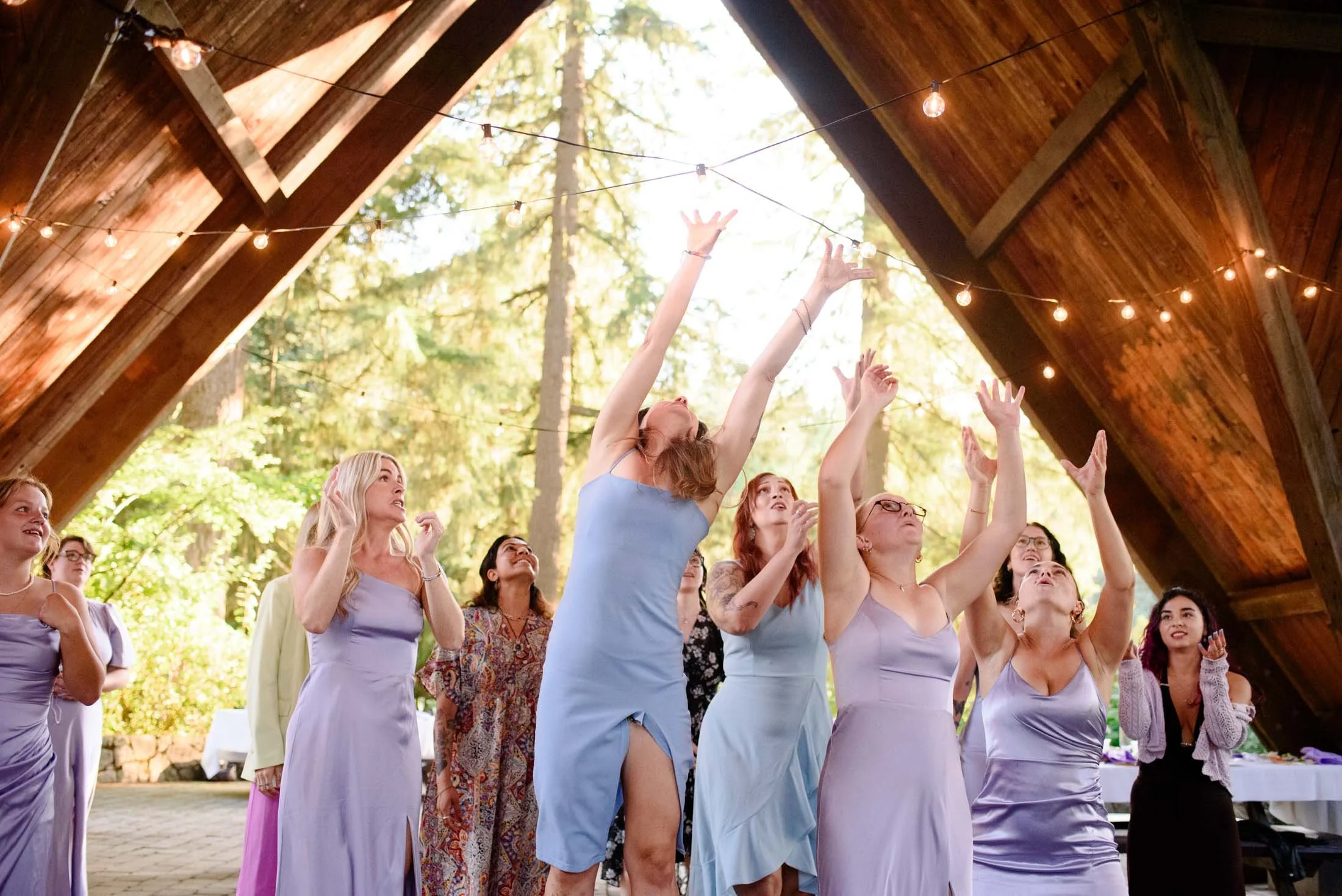 Wedding photograph at Portland's Hoyt Arboretum of Women in pastel dresses celebrating at a wedding under a wooden pavilion with string lights, reaching up joyfully.