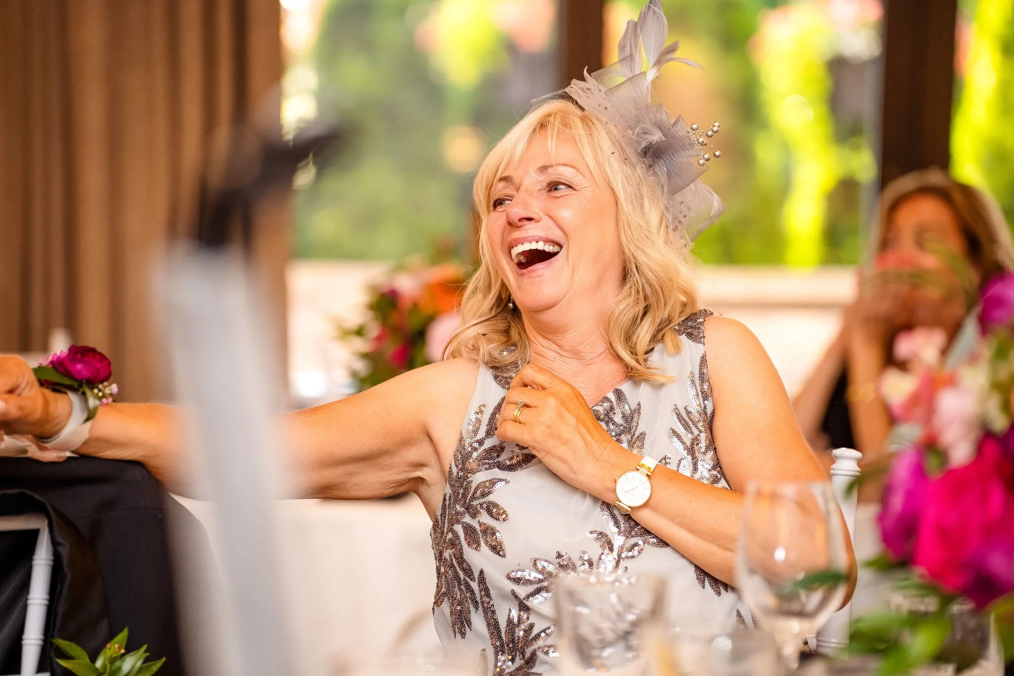 A woman in a decorated dress happily laughing at a celebration or party, with another woman in the background covering her mouth, surrounded by flowers and bright greenery at Abernethy Center Veiled Garden and Ballroom.