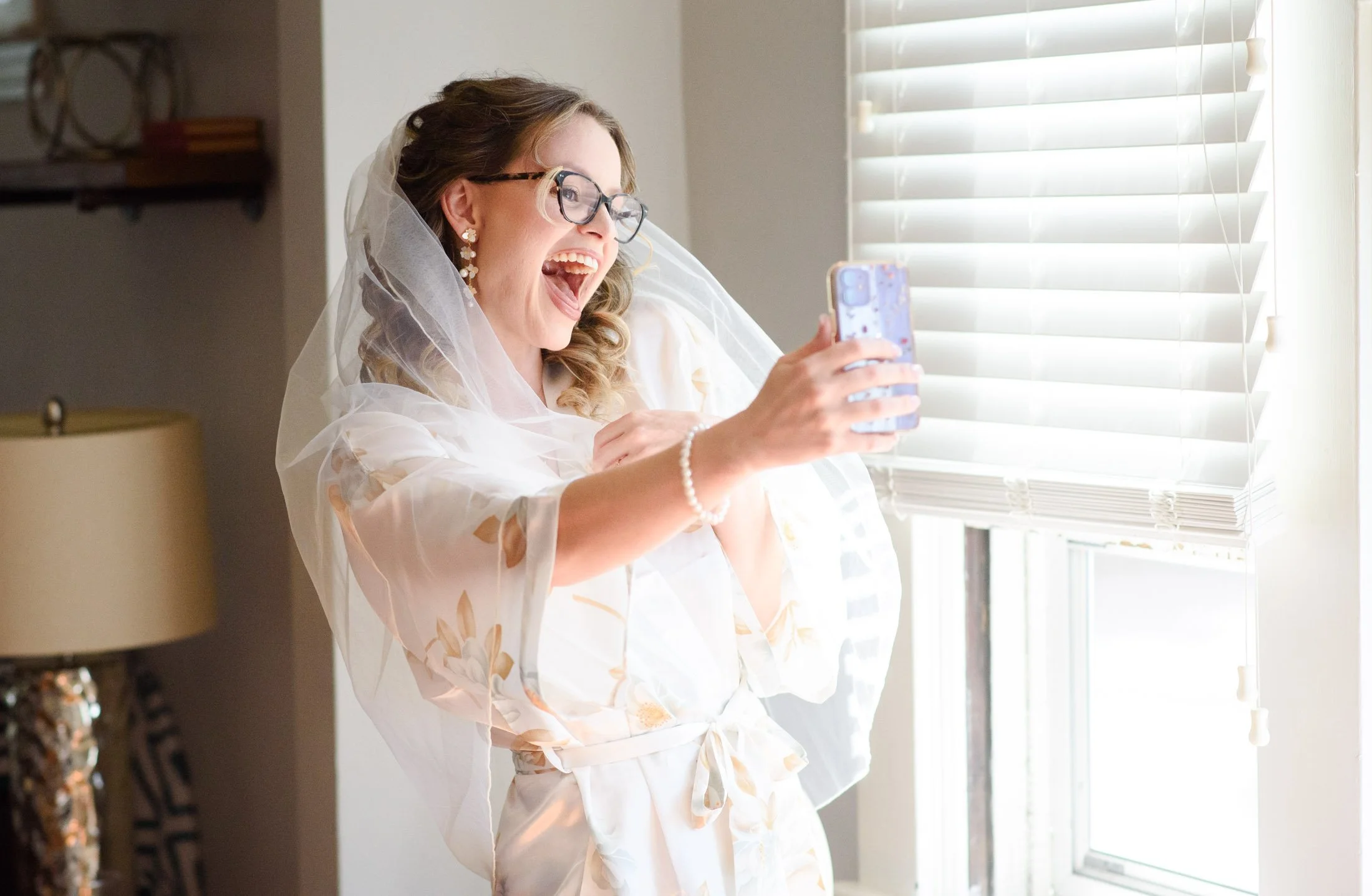 Wedding photo at Hoyt Arboretum of A woman in a bridal robe, wearing glasses and earrings, smiling and taking a selfie by a window with sunlight streaming in.