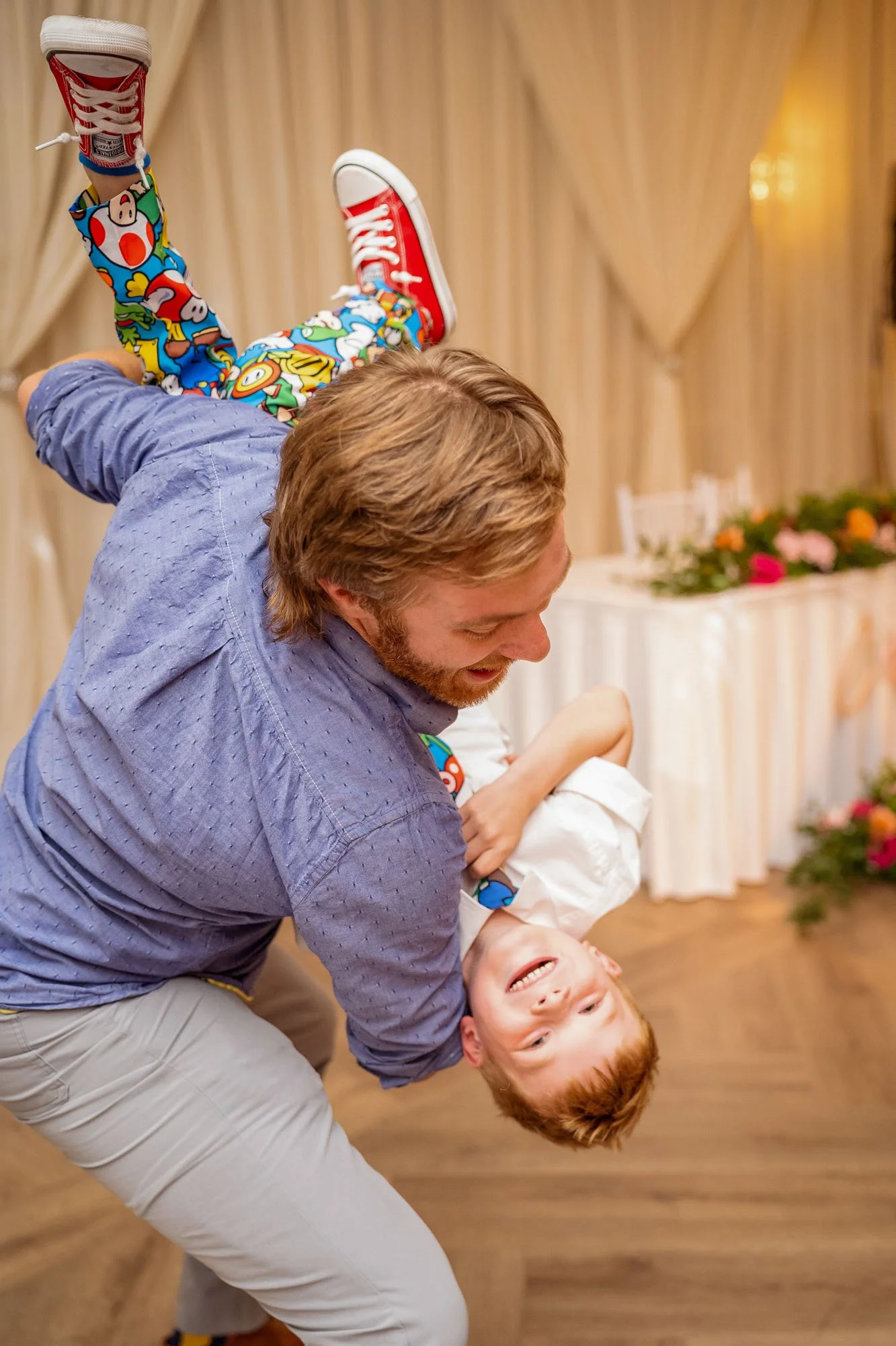 A man with light brown hair and a beard is playfully dancing with a smiling young boy with red hair, both in a warmly lit room with cream-colored curtains and floral decorations in the background at Abernethy Center Veiled Garden and Ballroom.