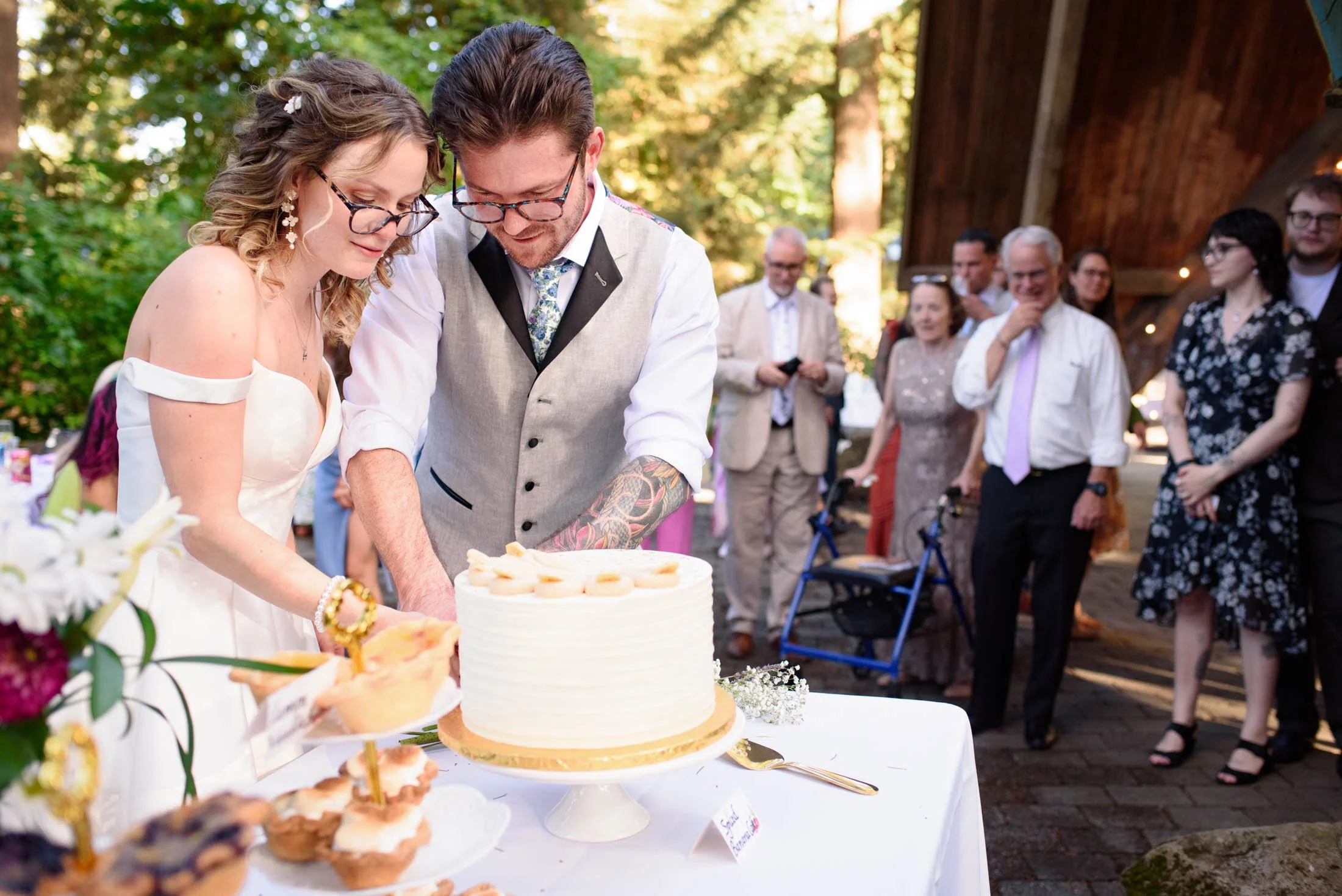 Wedding photograph at Portland's Hoyt Arboretum of Bride and groom cutting wedding cake outdoors with guests watching.