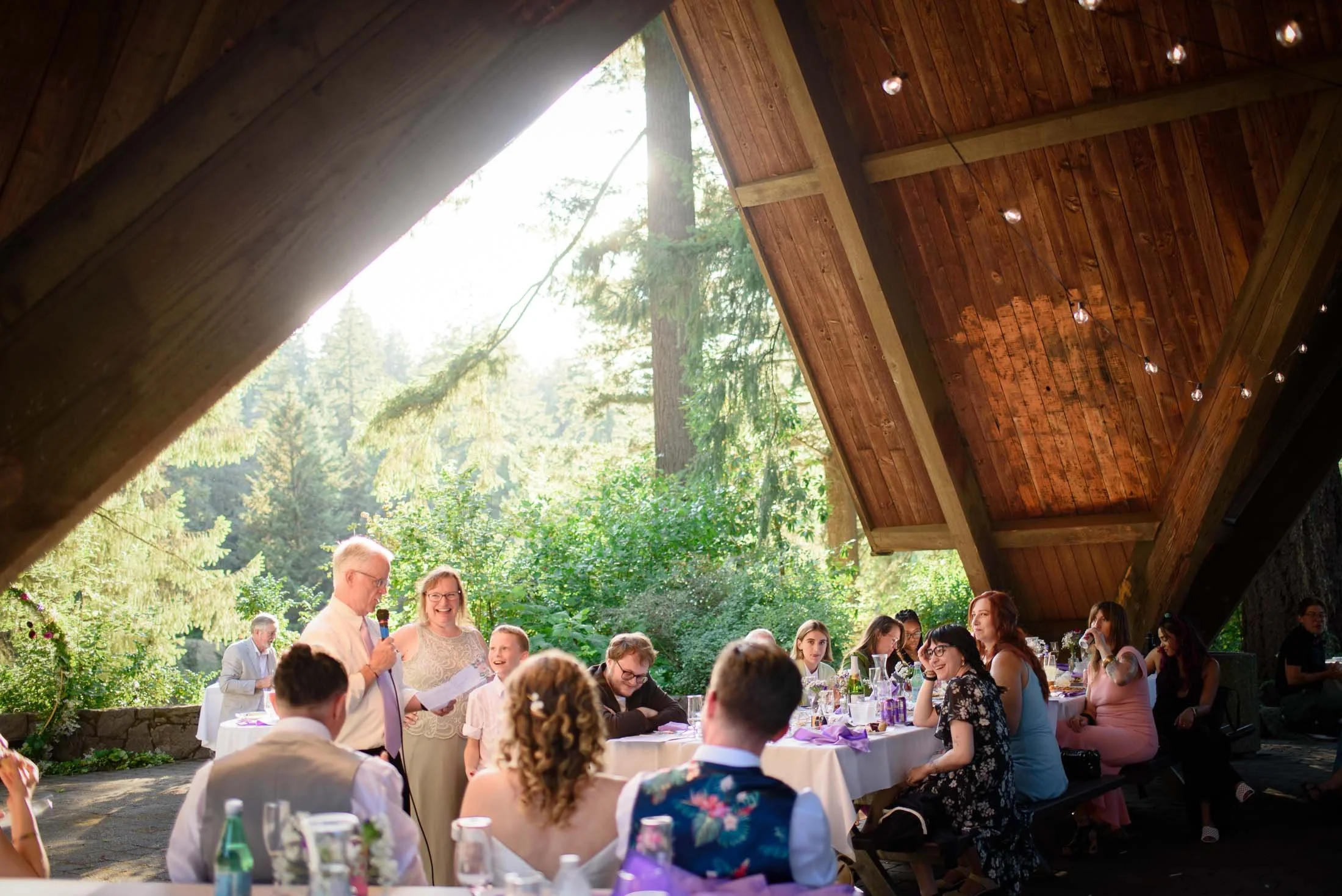 Wedding photograph at Portland's Hoyt Arboretum of People gathered at a wedding reception under a wooden pavilion with string lights, surrounded by greenery and tall trees, sunlight shining through.