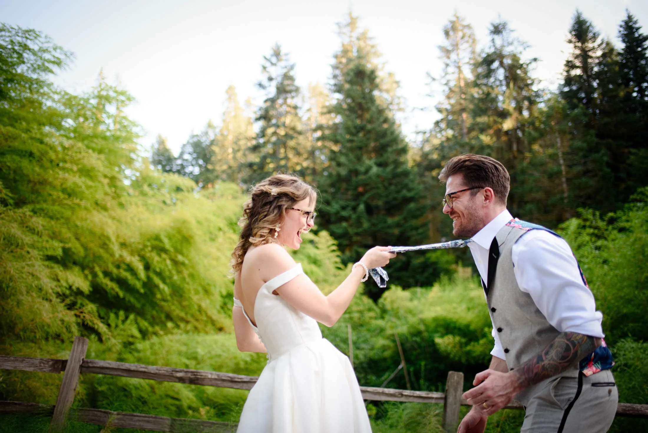 Wedding photograph at Portland's Hoyt Arboretum of A bride and groom laughing and playing with a tie at an outdoor wedding ceremony, surrounded by greenery and trees.