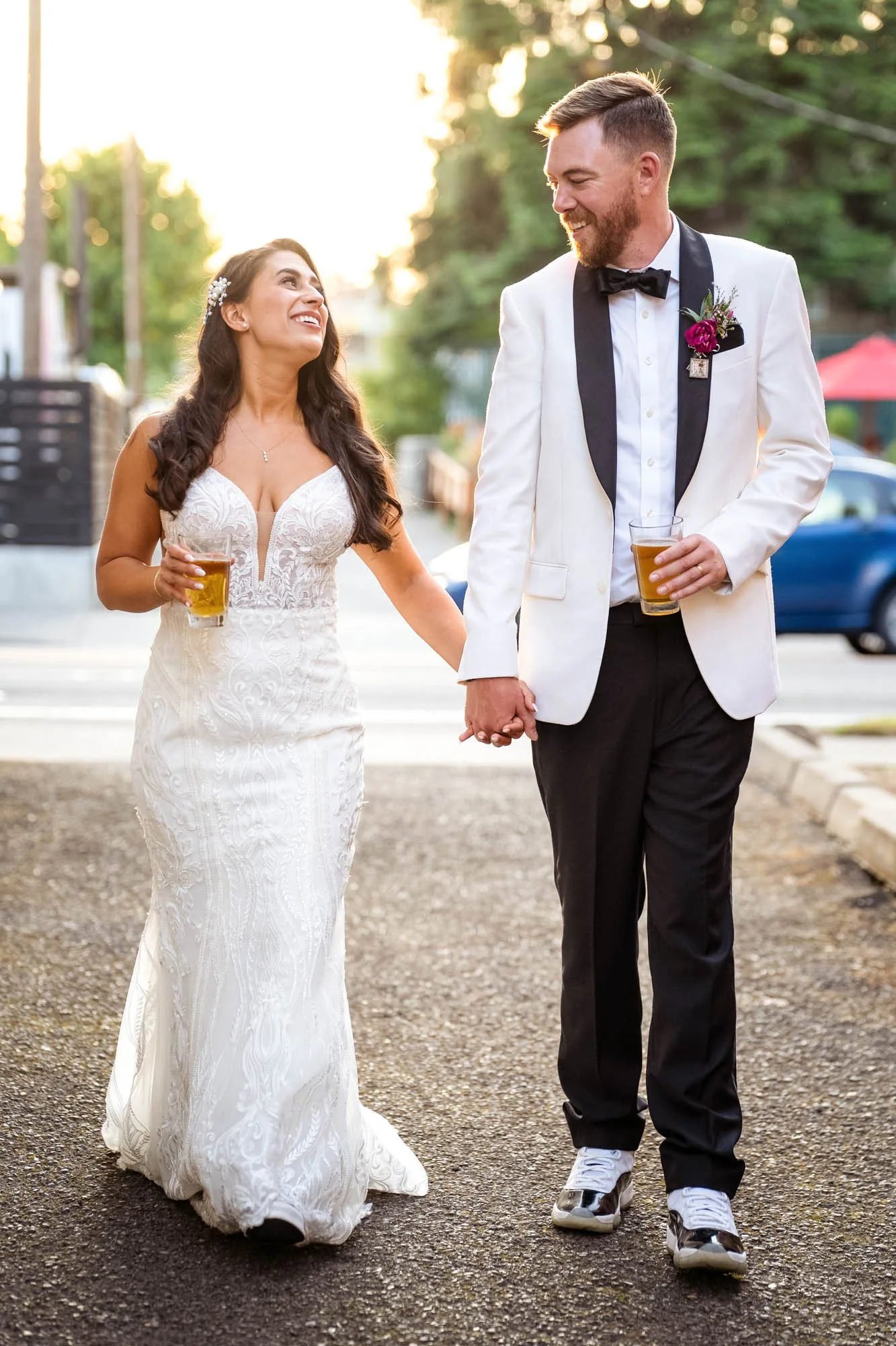 A bride and groom walking outdoors holding hands, both holding glasses of beer, smiling at each other, with sunlight and trees in the background at Abernethy Center Veiled Garden and Ballroom.
