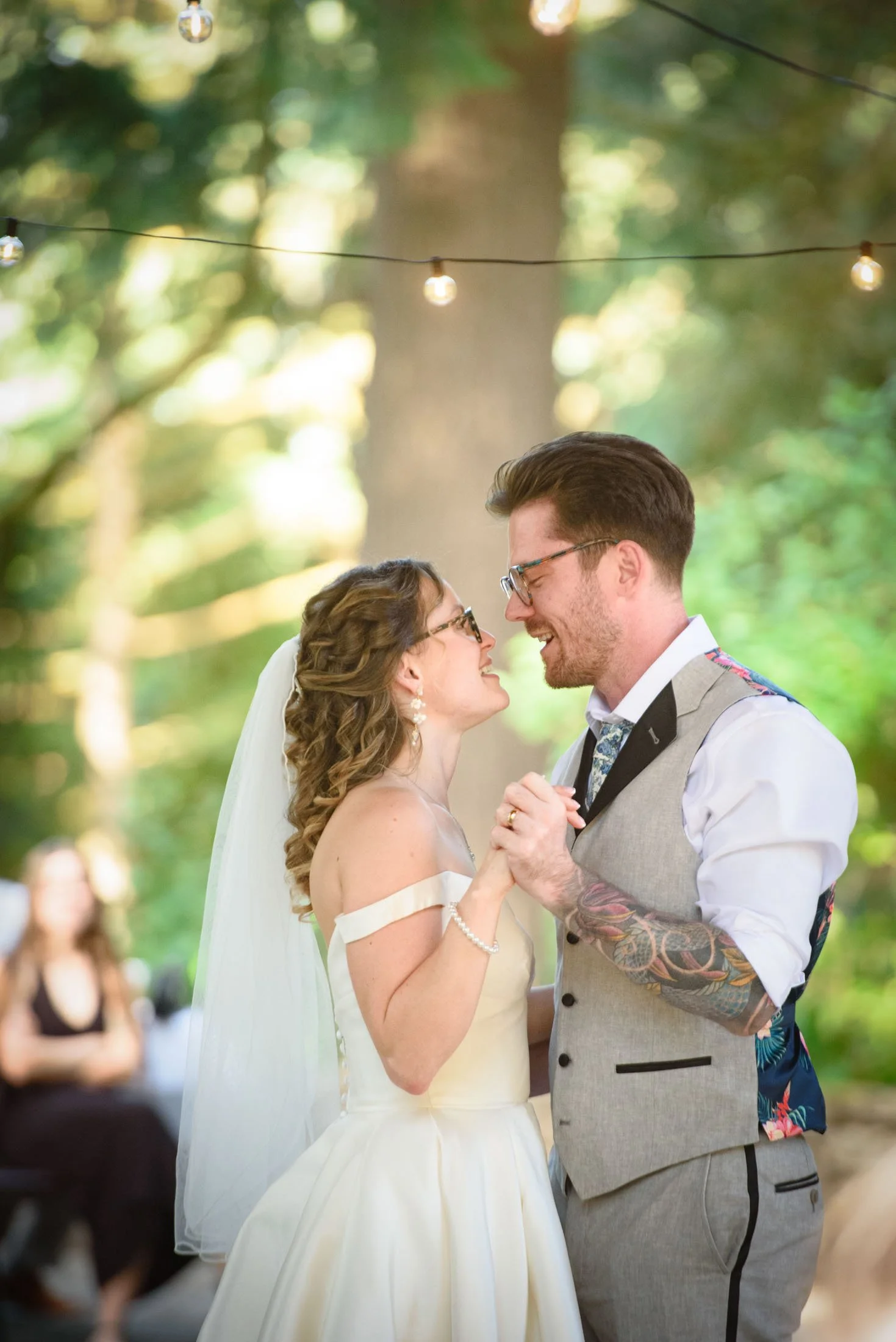 Wedding photograph at Portland's Hoyt Arboretum of A bride and groom sharing their first dance outdoors under string lights, surrounded by trees and blurred guests in the background.