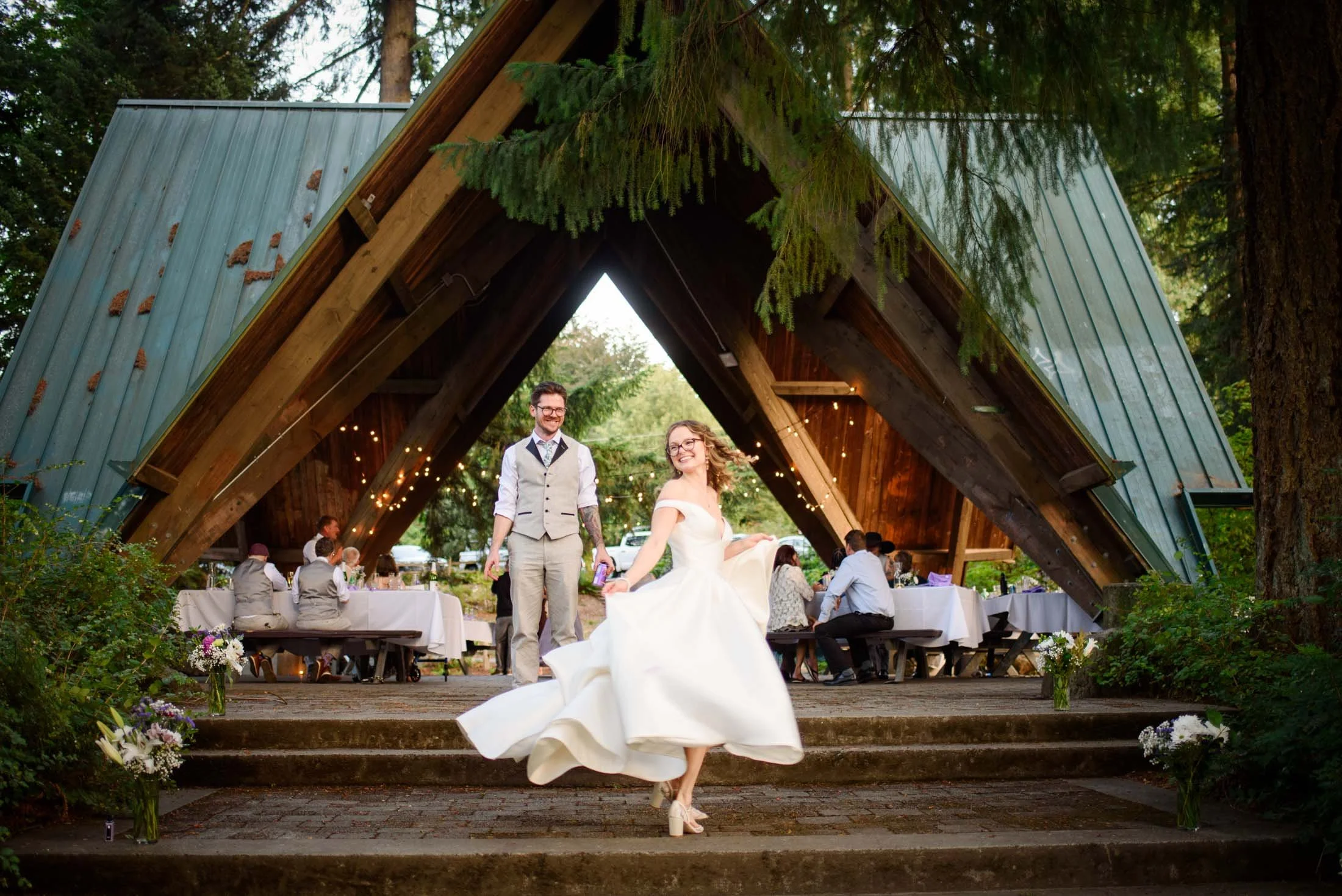 Wedding photograph at Portland's Hoyt Arboretum of Bride and groom dancing at their wedding reception under a wooden A-frame pavilion decorated with string lights, surrounded by guests seated at tables, with greenery and trees in the background.