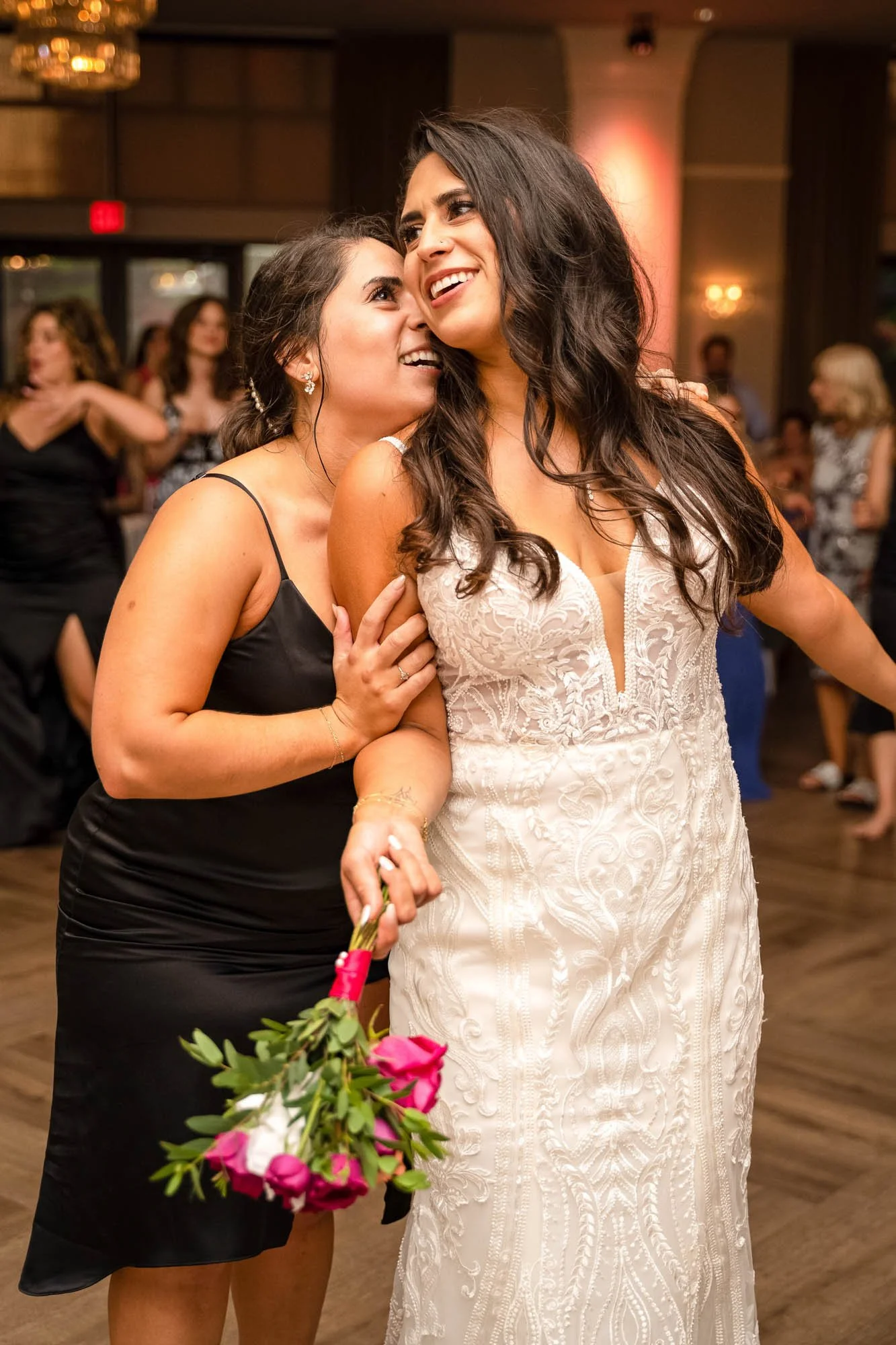 Wedding photograph at Abernethy Center Ballroom of two women at a celebration, one in a white wedding dress and the other in a black dress, sharing a joyful moment with the woman in the black dress holding a bouquet of pink and white flowers.