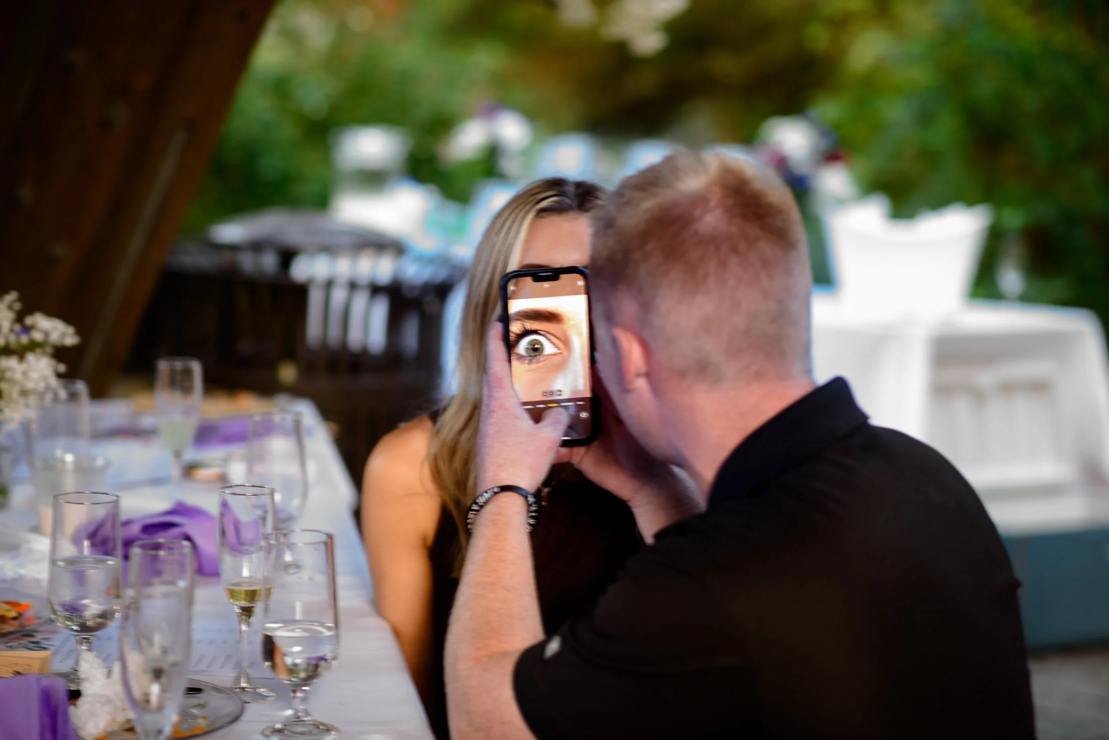 A man taking a close-up photo of a woman with his phone, with her face reflected in the screen, at a table set for a celebration or event outdoors.
