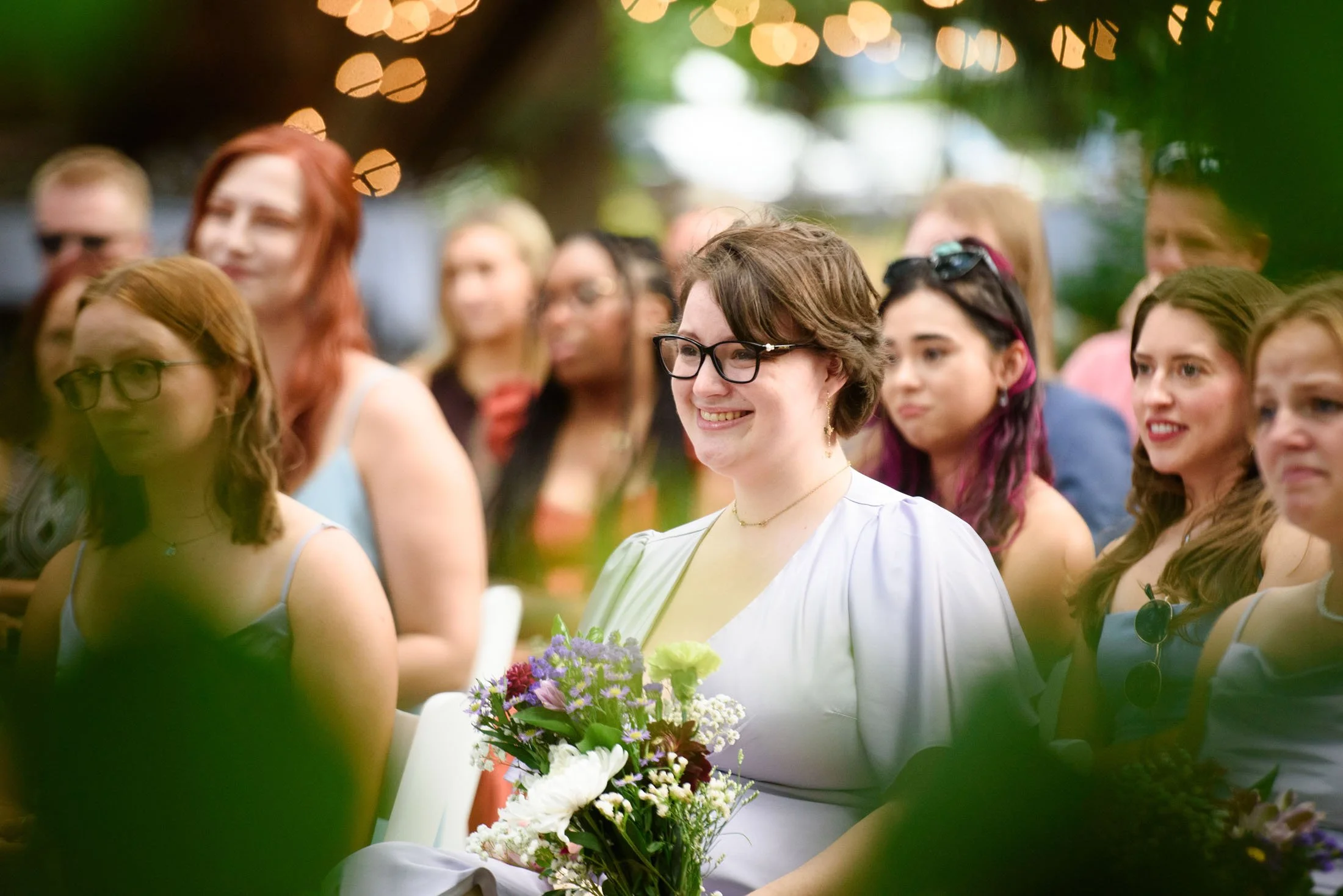 A group of women outdoors during daytime, dressed in casual summer clothing, with one woman in the center holding a bouquet of flowers. The woman in the center is smiling and wearing glasses, surrounded by other women who are also smiling or looking 