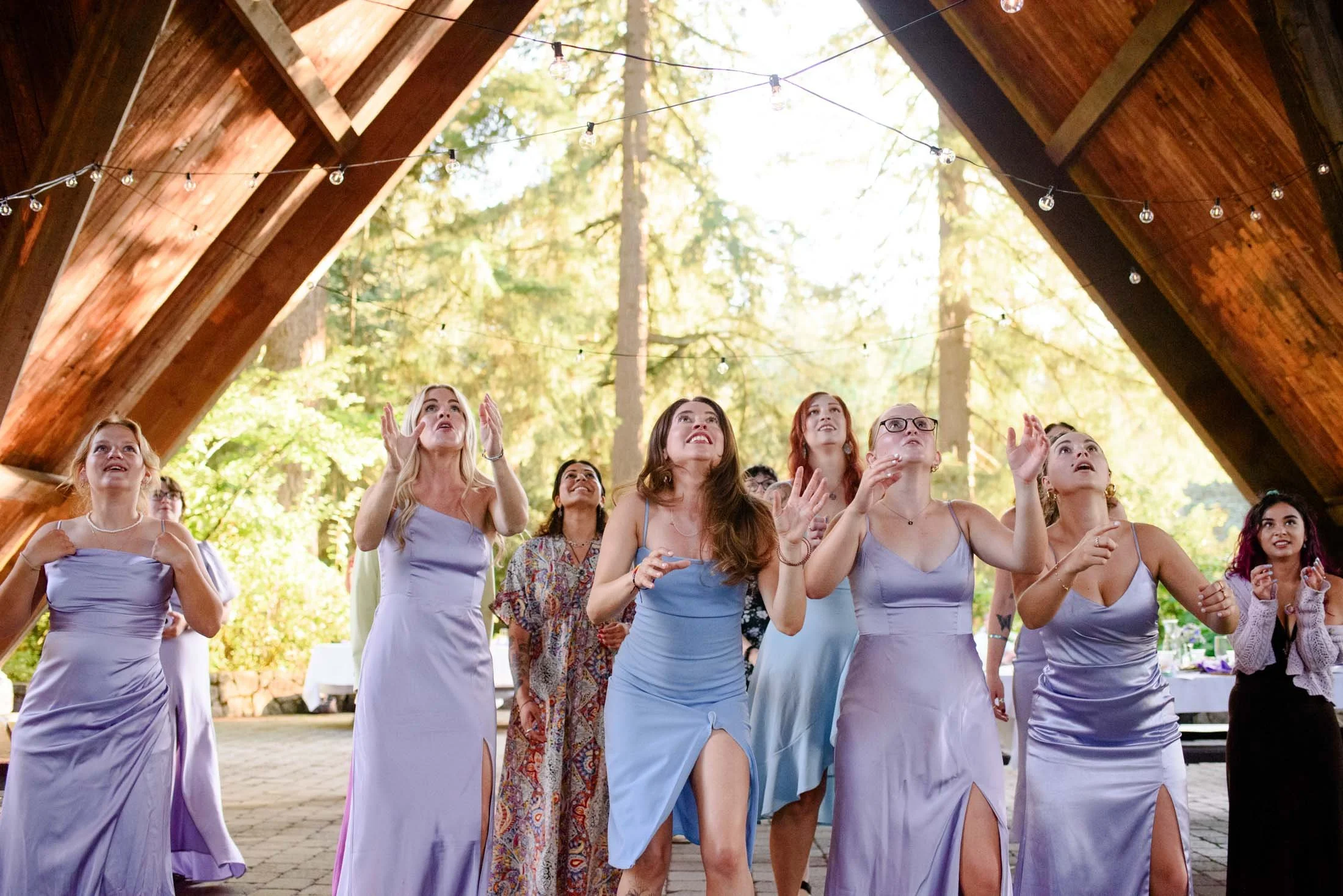 Wedding photograph at Portland's Hoyt Arboretum of Group of women in lavender dresses dancing at a wedding reception outdoors under a wooden pavilion with string lights and tall trees in the background.