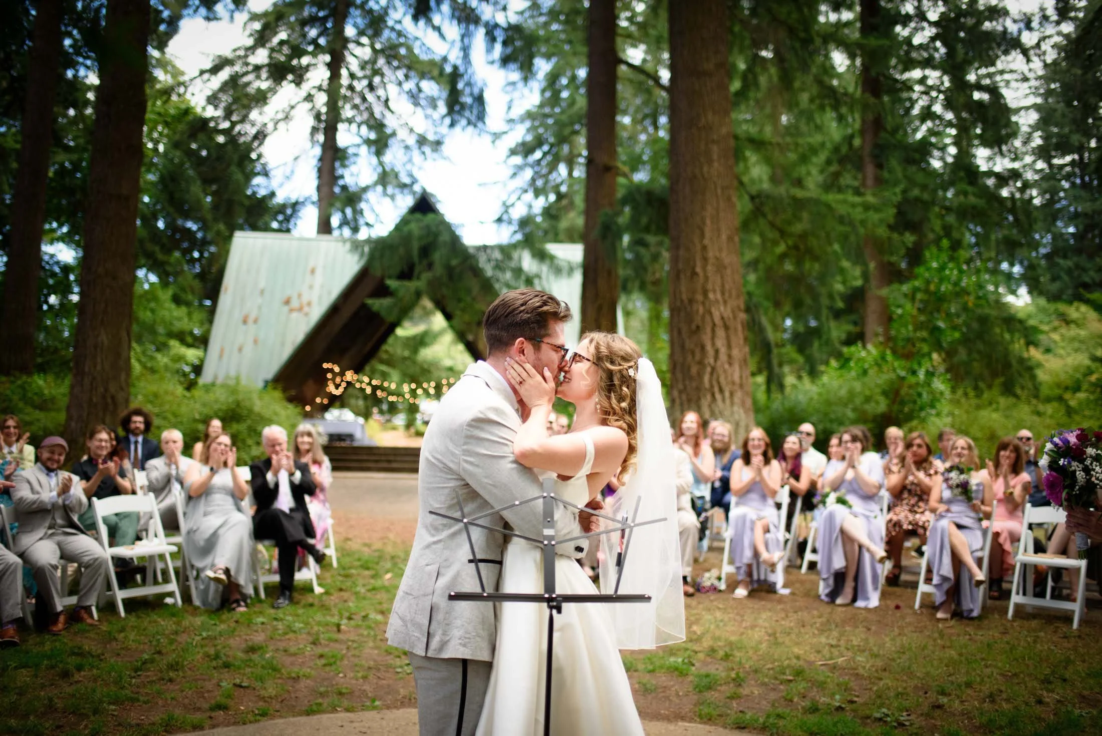 Wedding photo at Hoyt Arboretum of A couple sharing a kiss during a wedding ceremony outdoors, surrounded by friends and family seated on white chairs in a forest setting with tall trees and a wooden structure in the background.