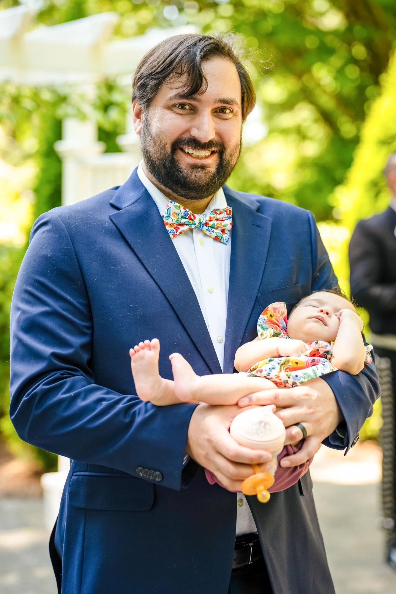 Wedding photo at Abernethy Center's Abernethy Chapel of A man with a beard in a blue suit and colorful bowtie holding a sleeping baby girl dressed in a patterned outfit outdoors with greenery in the background.