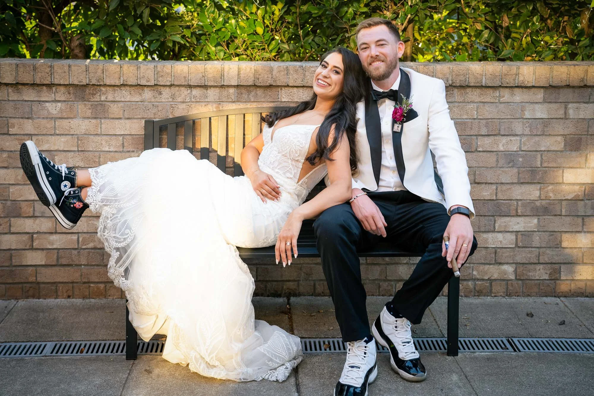 A bride and groom sitting on a black bench against a brick wall and greenery background, smiling  at Abernethy Center Veiled Garden and Ballroom.
