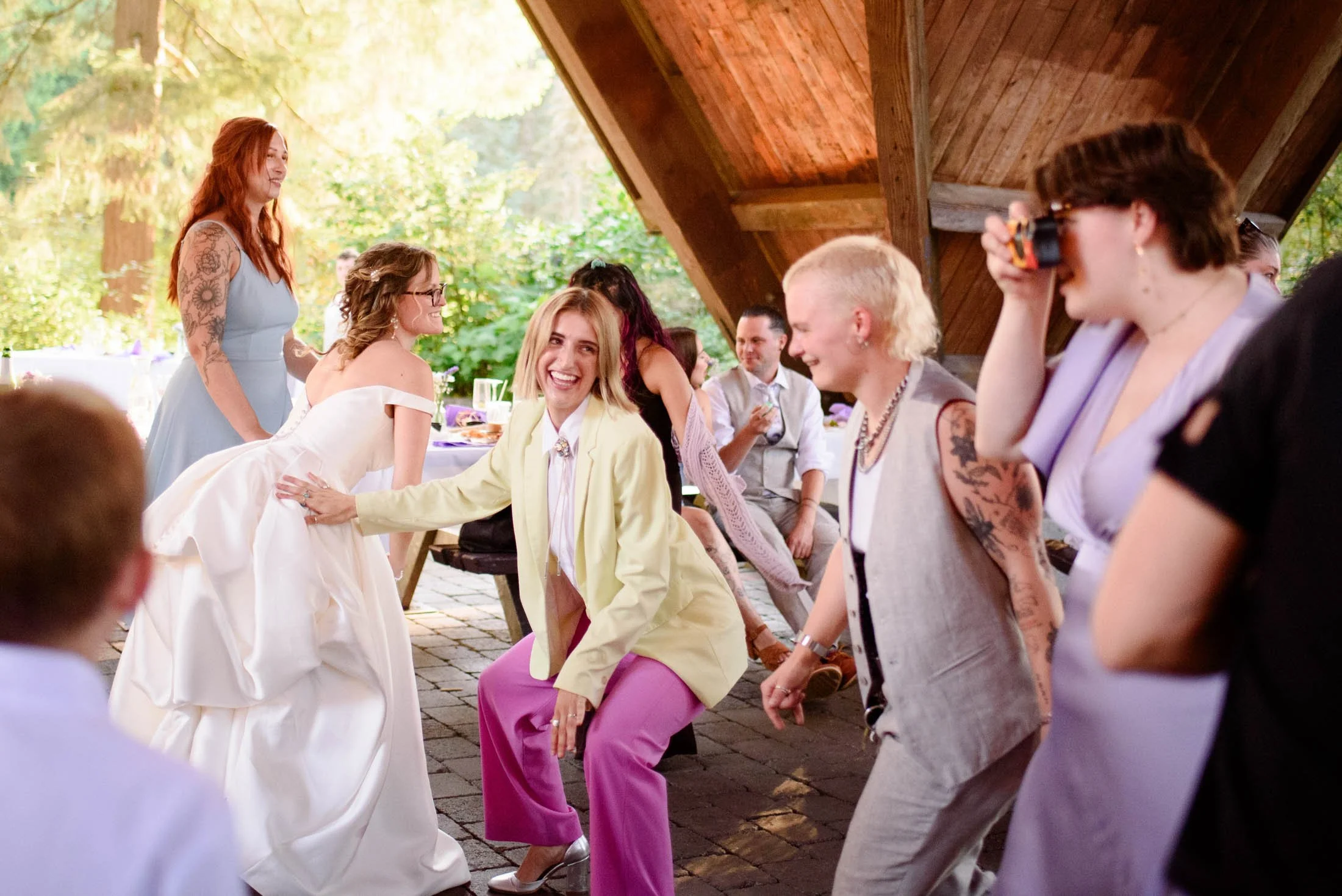 Wedding photograph at Portland's Hoyt Arboretum of Group of women dancing and laughing at an outdoor celebration or wedding reception, with greenery and a wooden structure in the background.