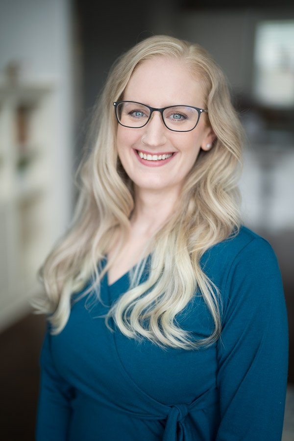Portrait of a woman with long blonde hair, wearing glasses and a blue top, smiling in an indoor setting.