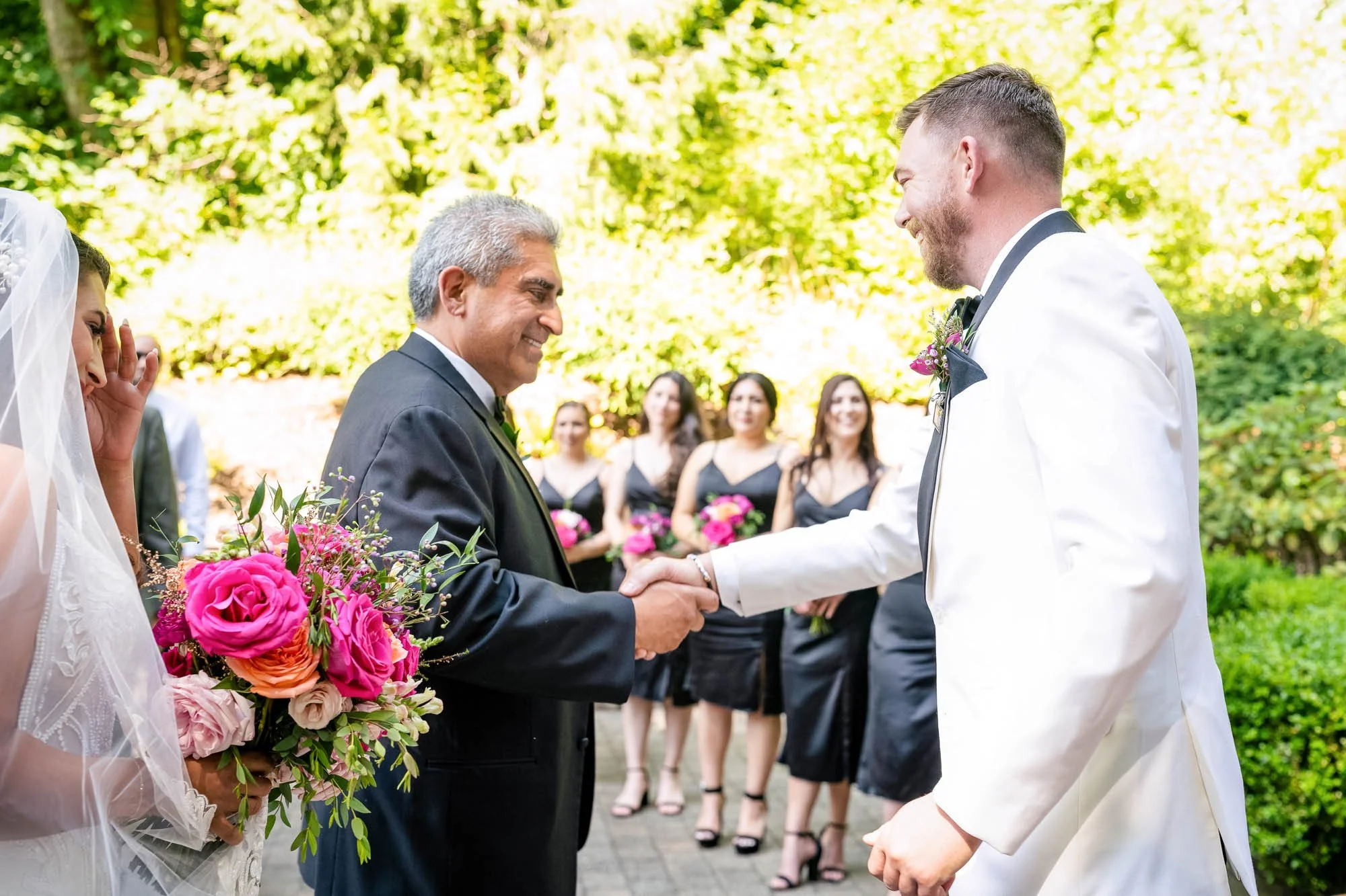 Wedding photography at Abernethy Center of A wedding ceremony outdoors with a bride holding a bouquet of pink and peach flowers, two men shaking hands, and bridesmaids in black dresses holding pink bouquets in the background.