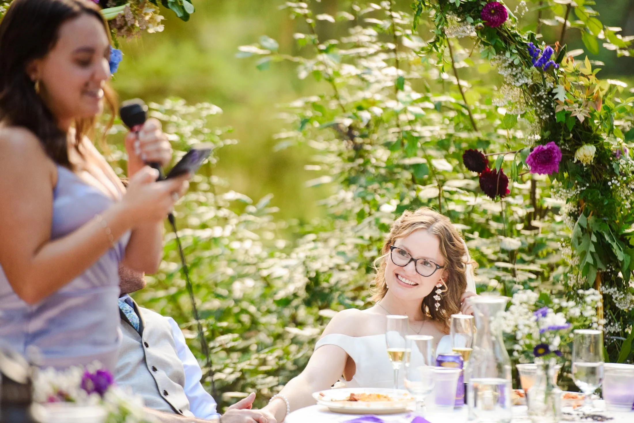 A smiling woman with glasses and earrings sitting at a table decorated with flowers and glassware during an outdoor wedding  at Hoyt Arboretum in Portland.