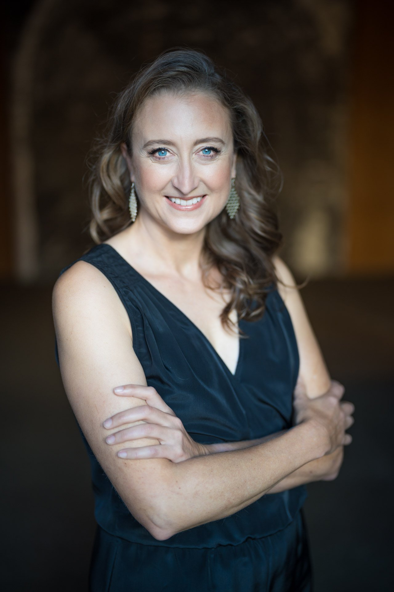 Portrait of a woman with long wavy brown hair, blue eyes, and earrings, wearing a black sleeveless dress, smiling with arms crossed, standing in a dimly lit background.