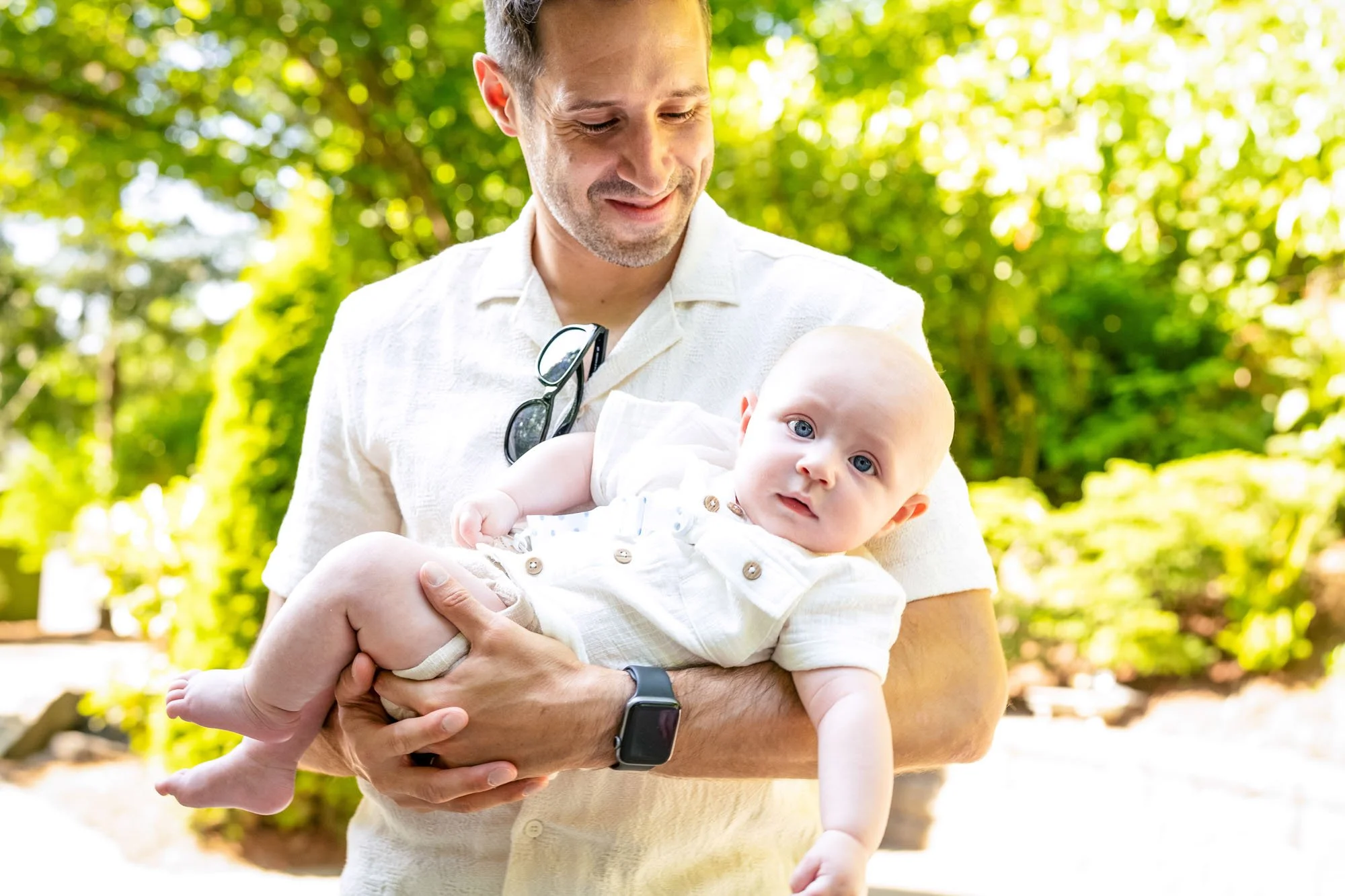 A man with a beard, wearing a white short-sleeve shirt and a smartwatch, holding a baby boy with blue eyes and a light complexion outdoors in a green park. The man has sunglasses hanging from his shirt collar and is smiling while the baby gazes at th