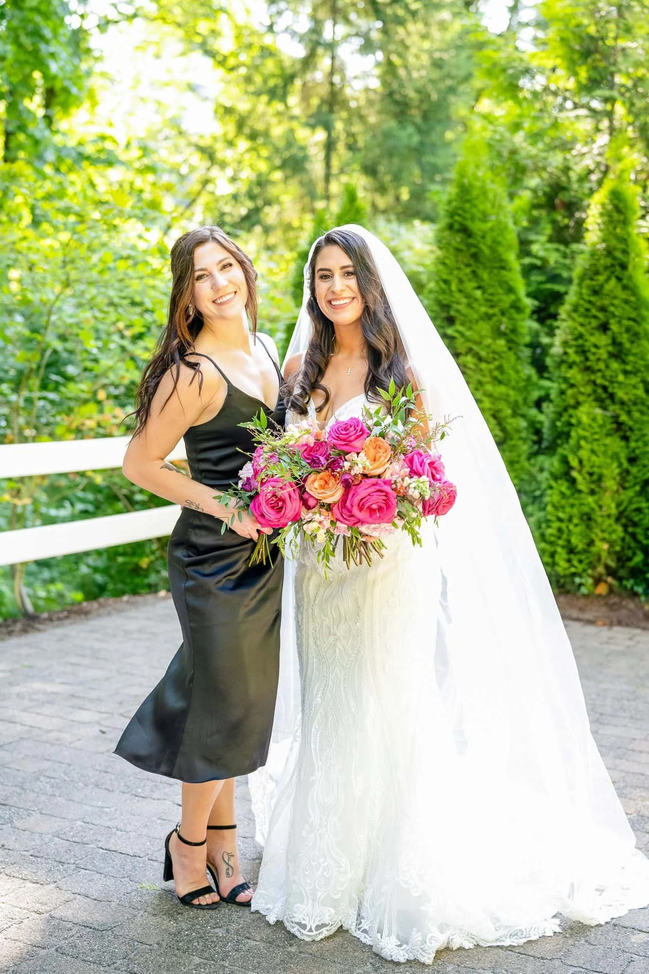 Wedding photo at Abernethy Center's Abigail's Two women, one in a white wedding dress with veil holding a large bouquet of pink and orange roses, and the other in a black dress, standing outdoors on pavement with greenery and trees in the background.